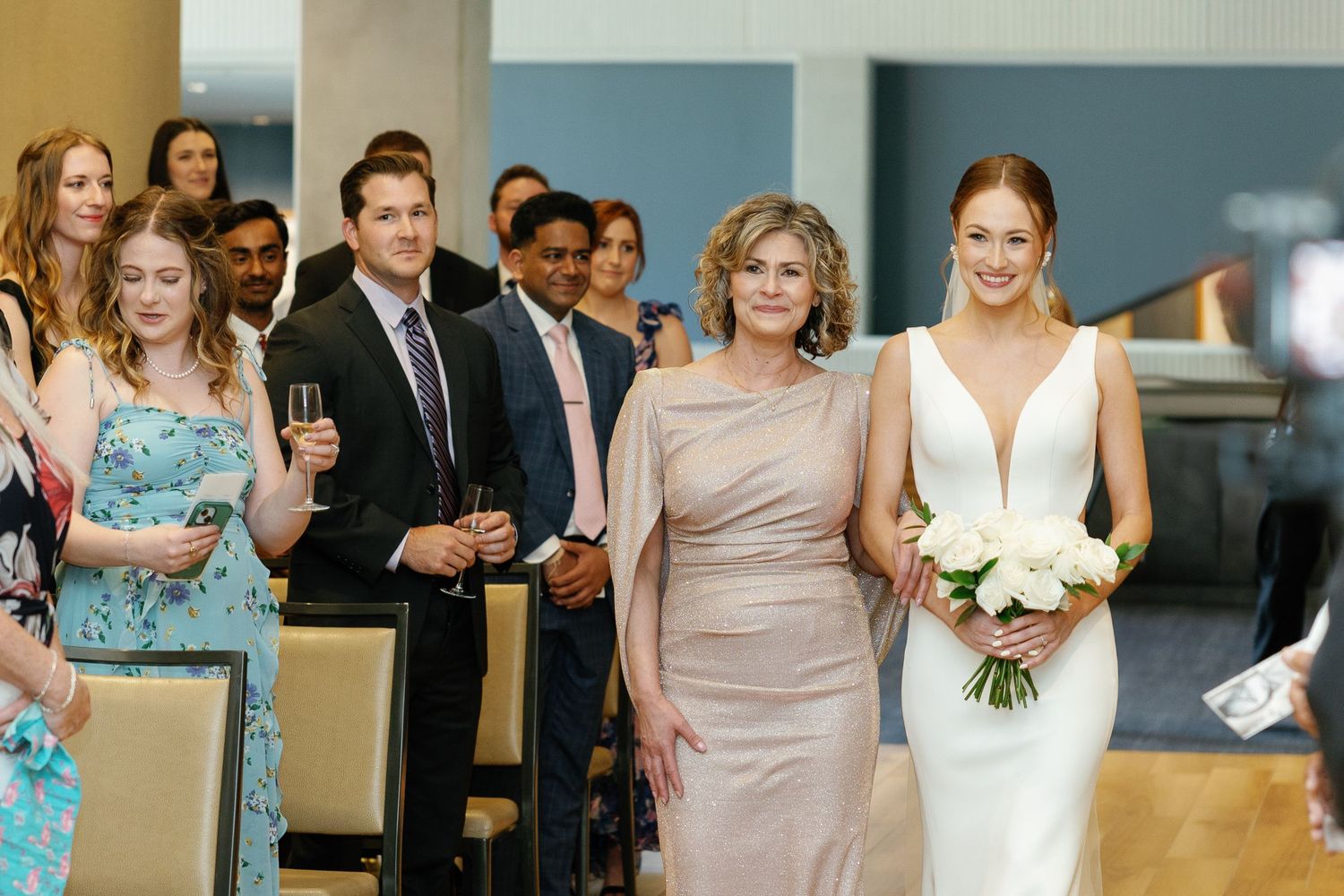 Wedding ceremony attendees gather in a modern venue with people wearing formal attire including teal and beige dresses.