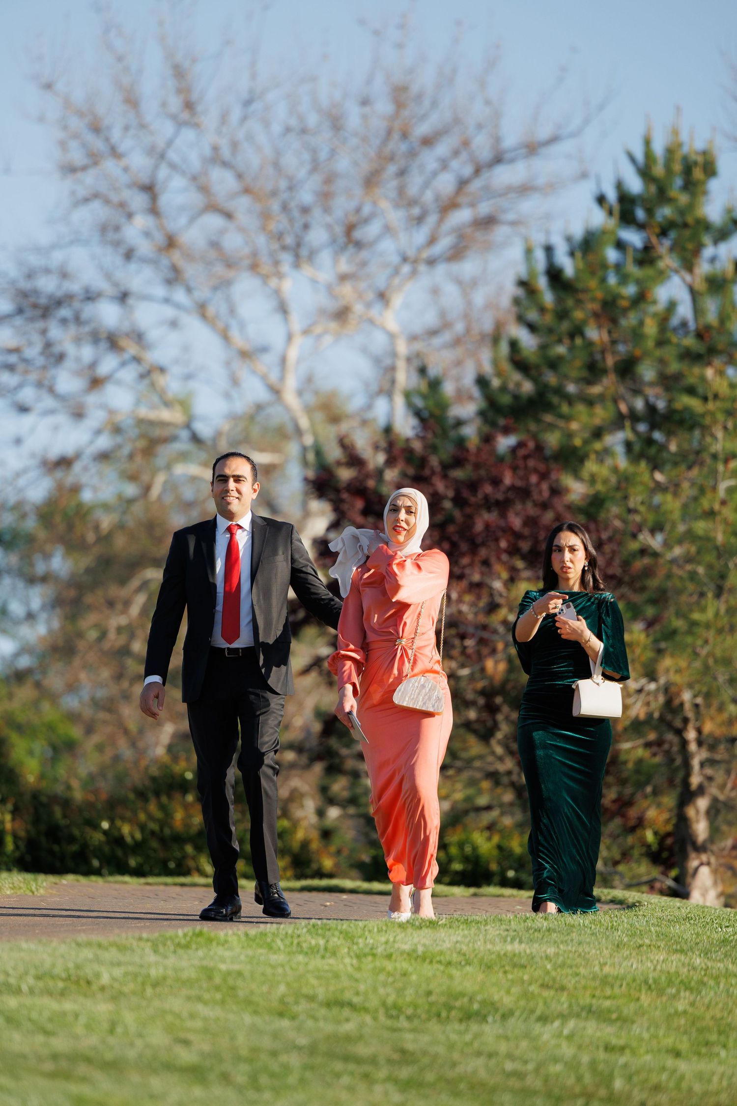 Three people in formal attire stand on a grassy hill outdoors during autumn.