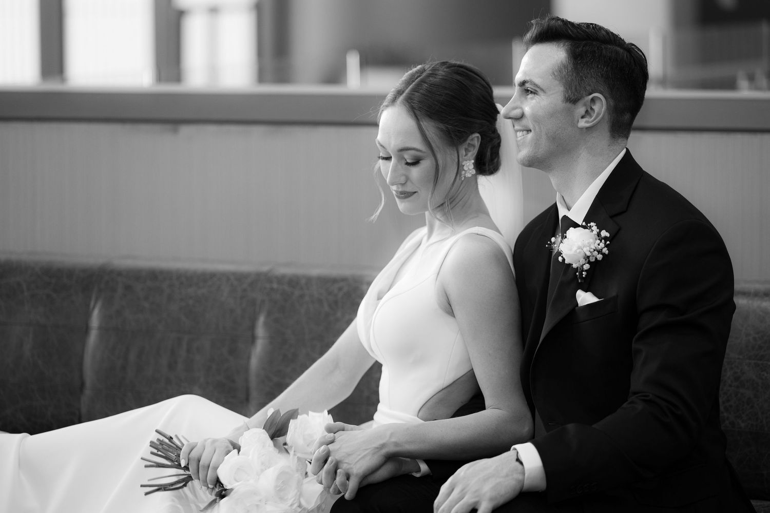 A bride and groom sit together in formal wedding attire during an intimate black and white moment.