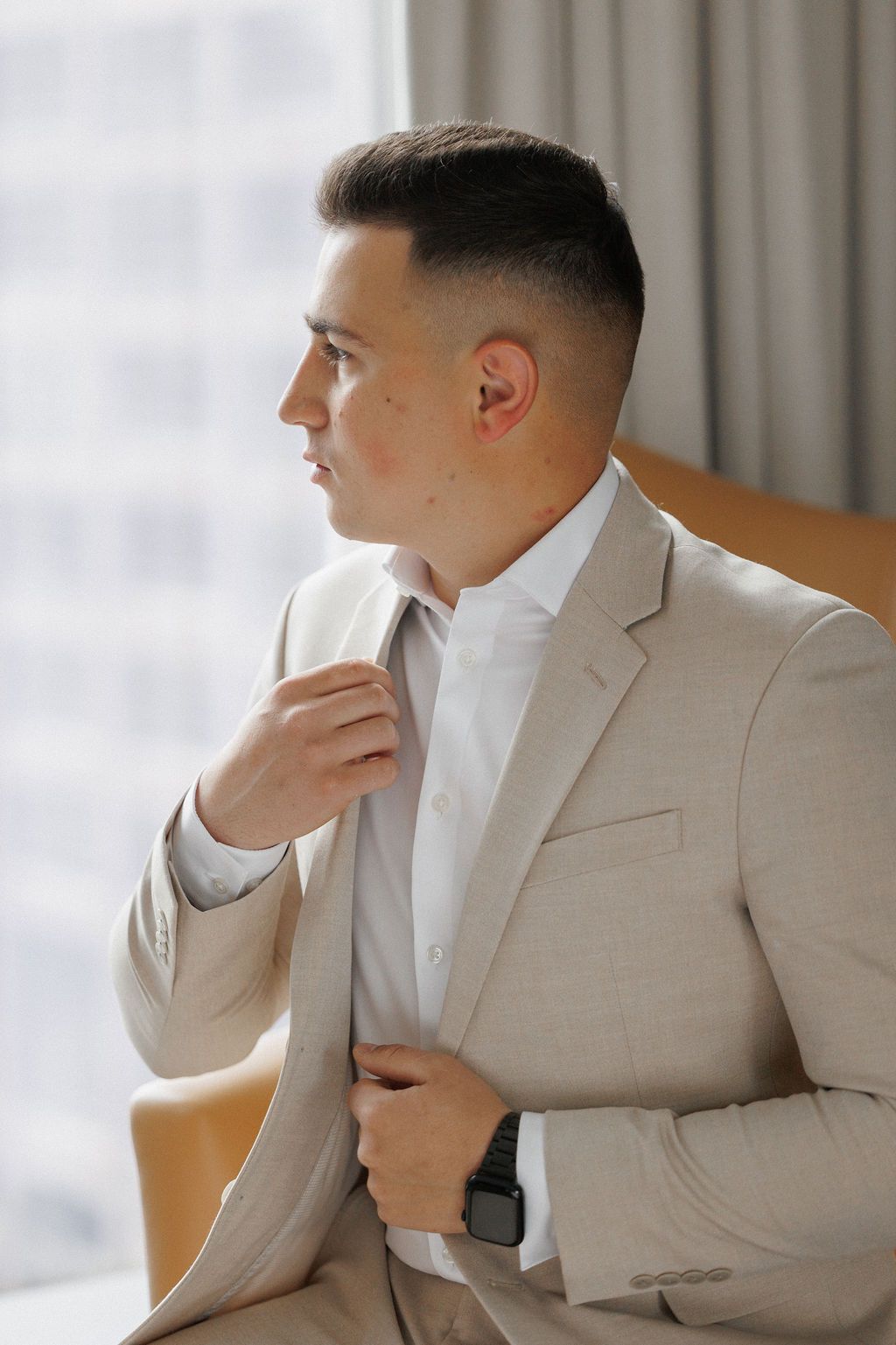 A person in a beige suit adjusts their collar while looking to the side in a bright interior setting.