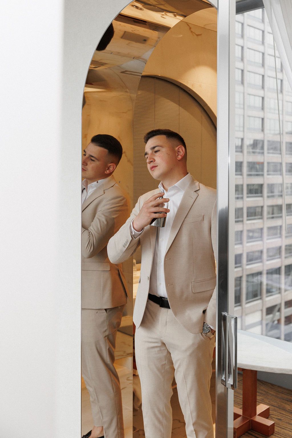 A person in a beige suit stands in front of a mirror in a modern high-rise building.