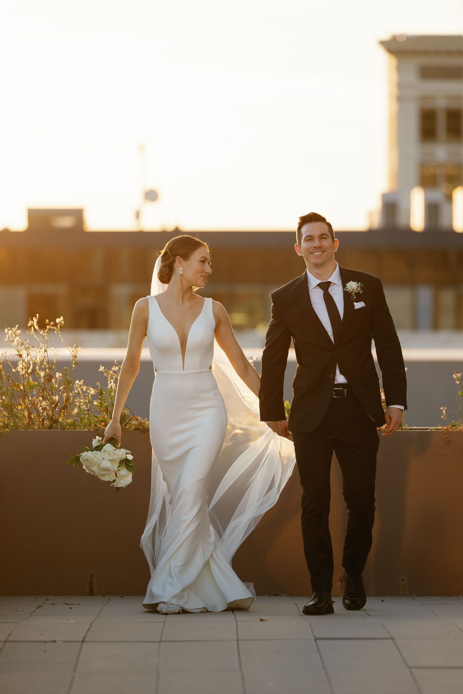 Elegant sunset wedding photo of couple walking together on rooftop with warm golden light.