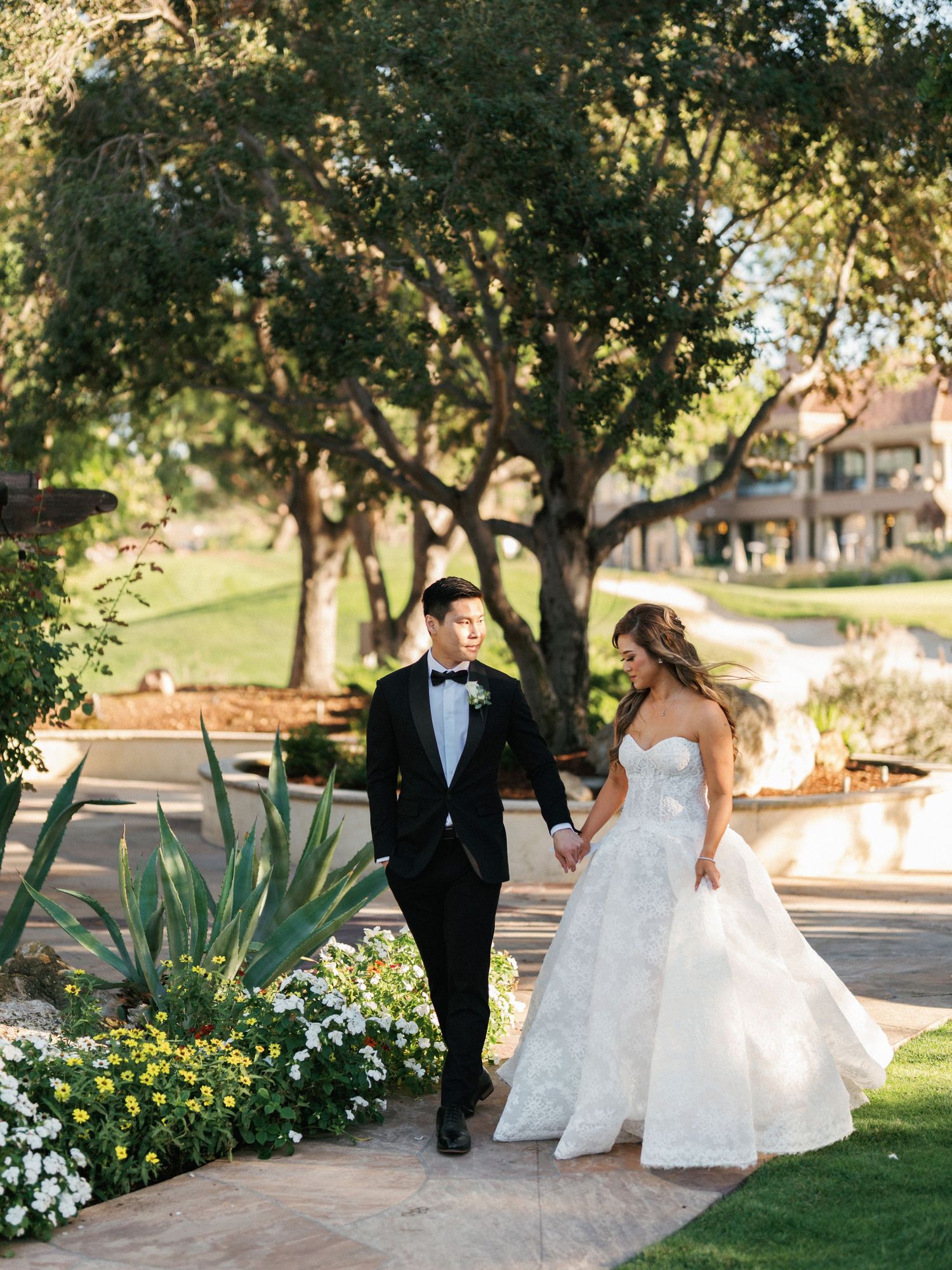 A couple walks hand in hand through a garden path with blooming flowers and trees.
