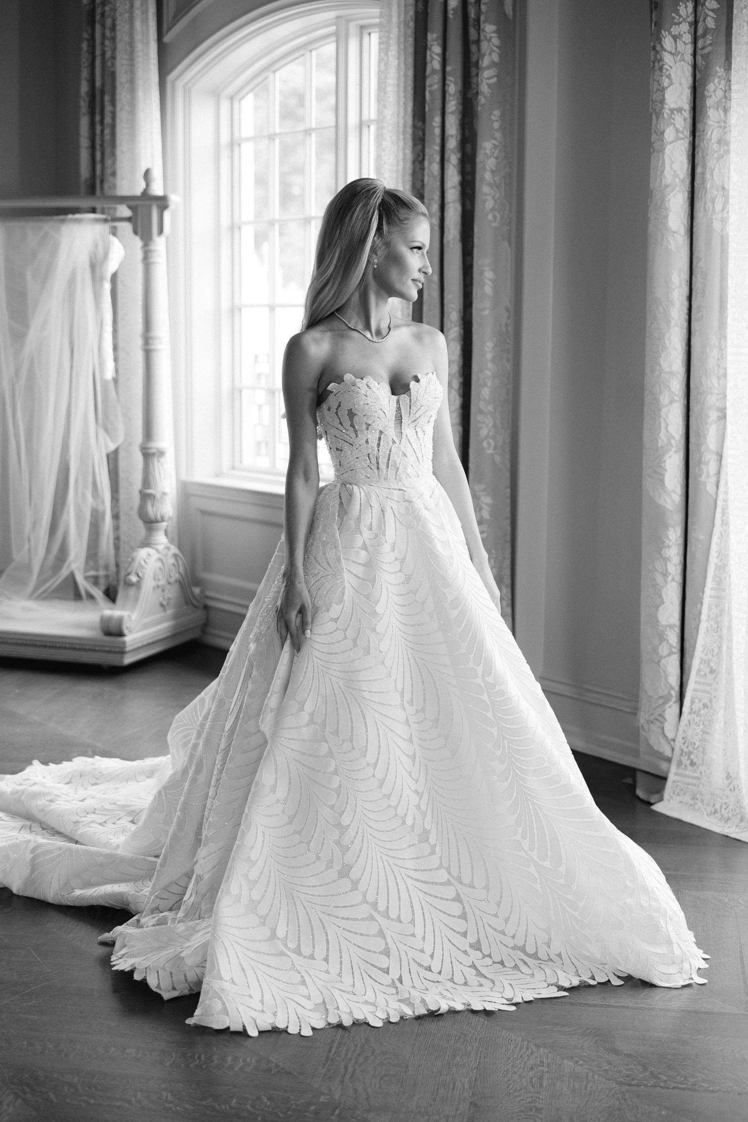 Bride in strapless feather-lace ballgown wedding dress standing by window in elegant room, black and white photo.