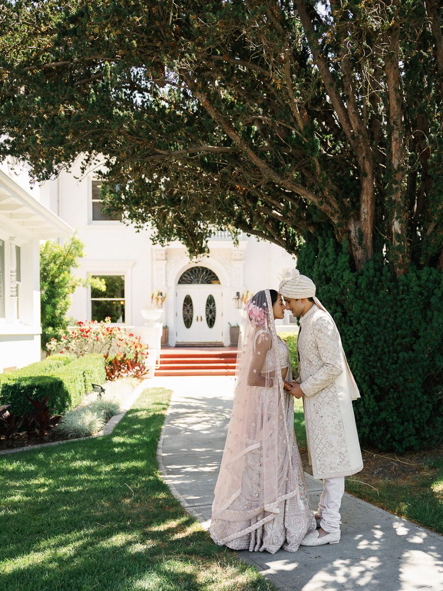 Wedding couple in traditional white attire walking together on a garden path with trees.