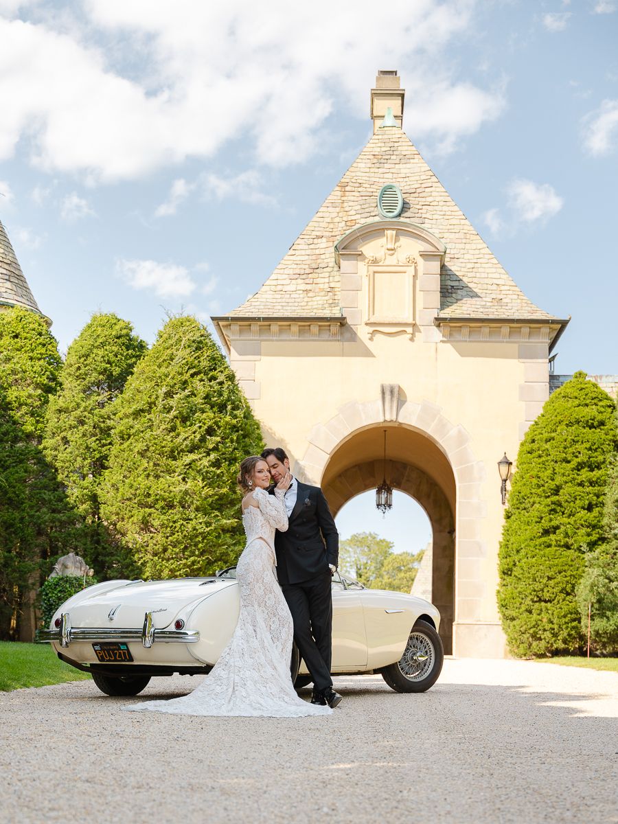 Classic silver sports car parked beside stone archway during wedding celebration with couple embracing in background.