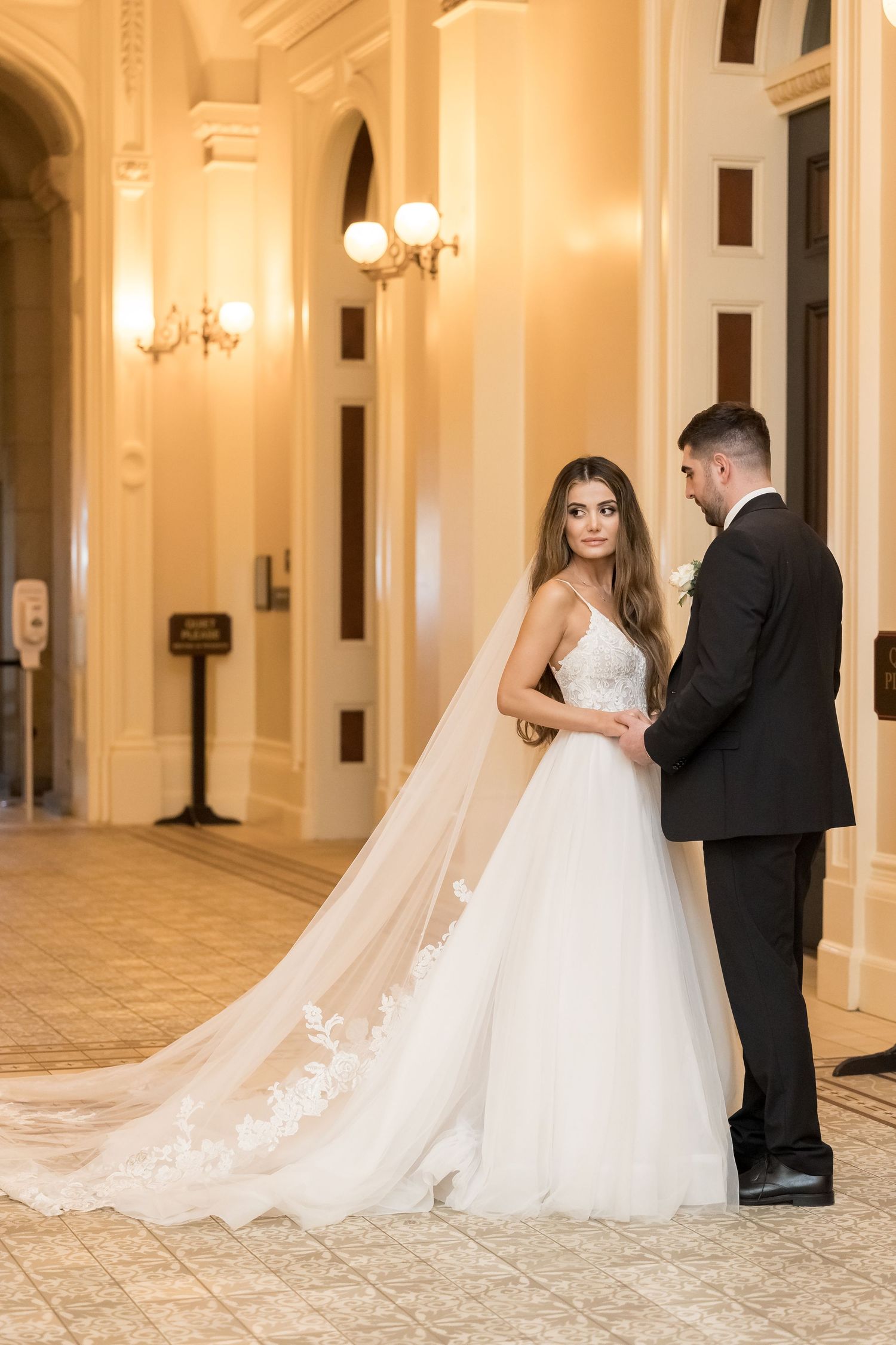 Elegant wedding couple poses in ornate hallway with golden lighting and classical architecture.