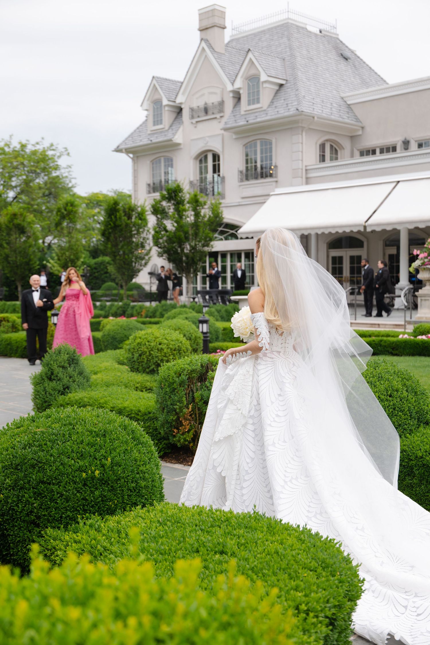 Bride in white lace gown and veil walks through manicured garden toward elegant estate venue.