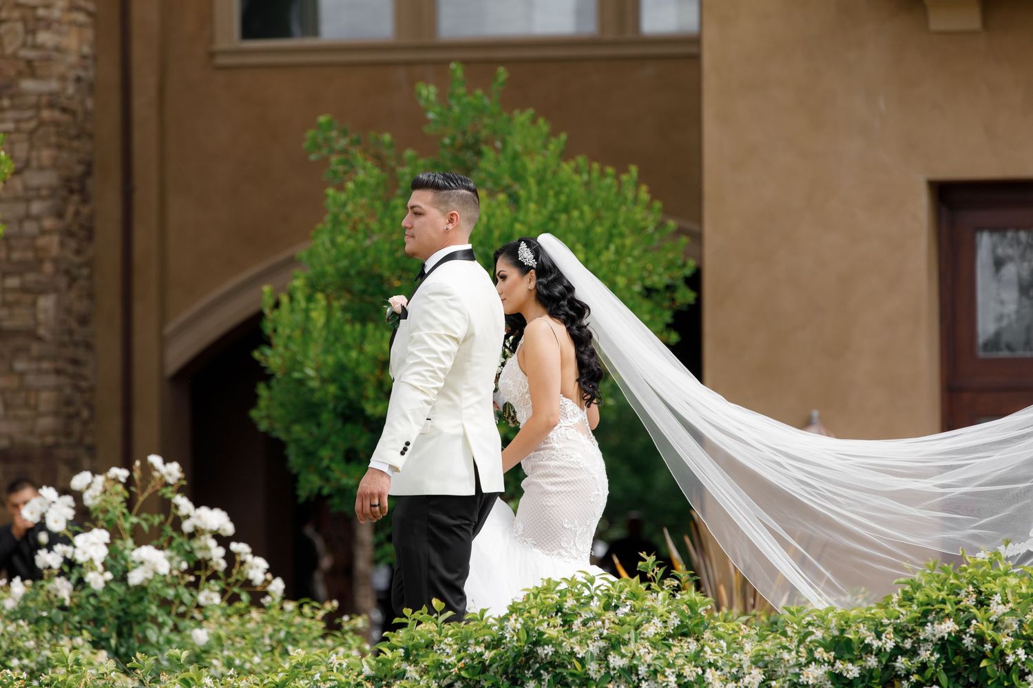 A bride in a flowing white gown with long veil stands back-to-back with a groom in white uniform at an elegant venue.