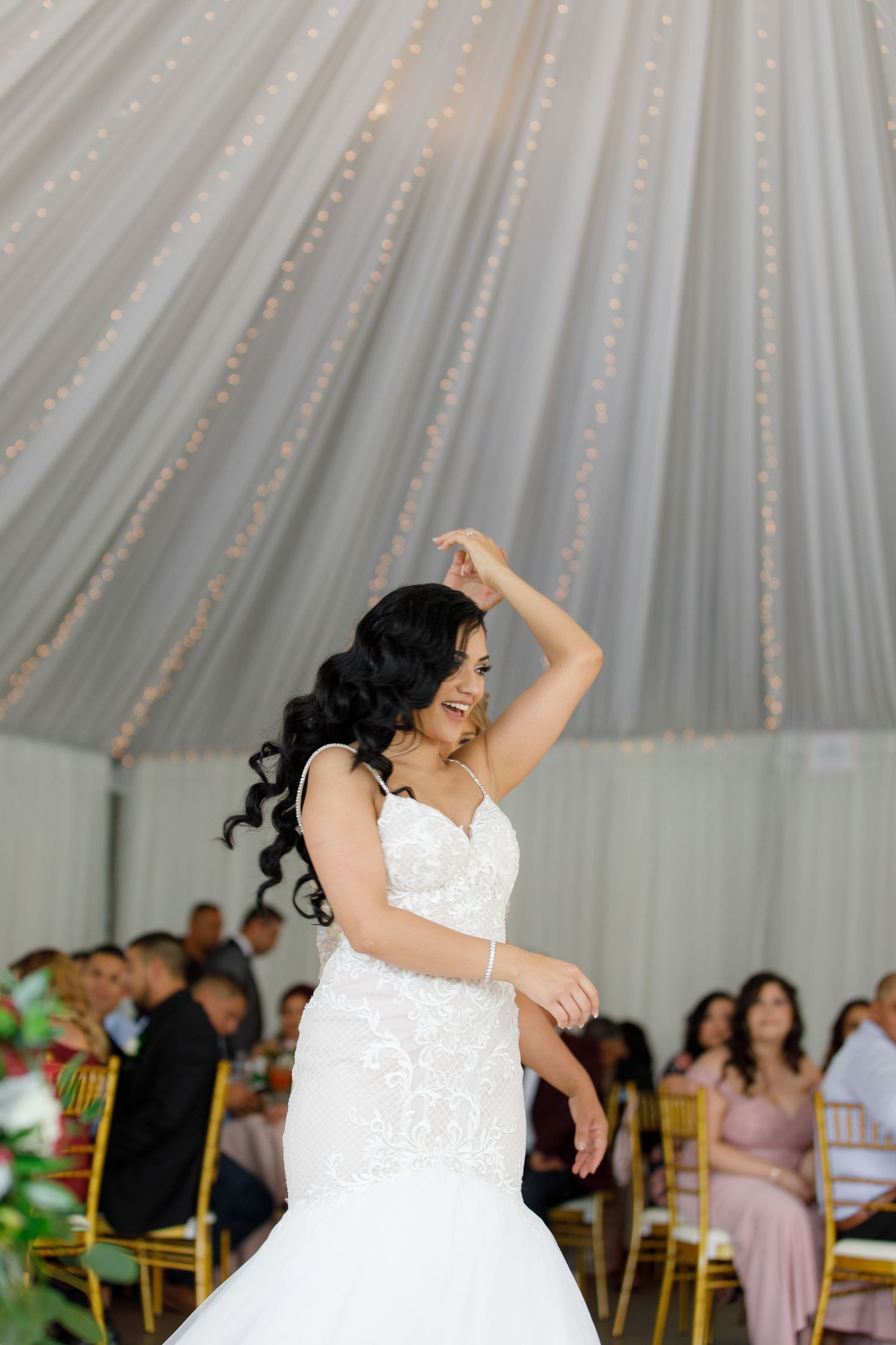 A person in a white wedding dress dances joyfully under a tent with string lights and guests in the background.