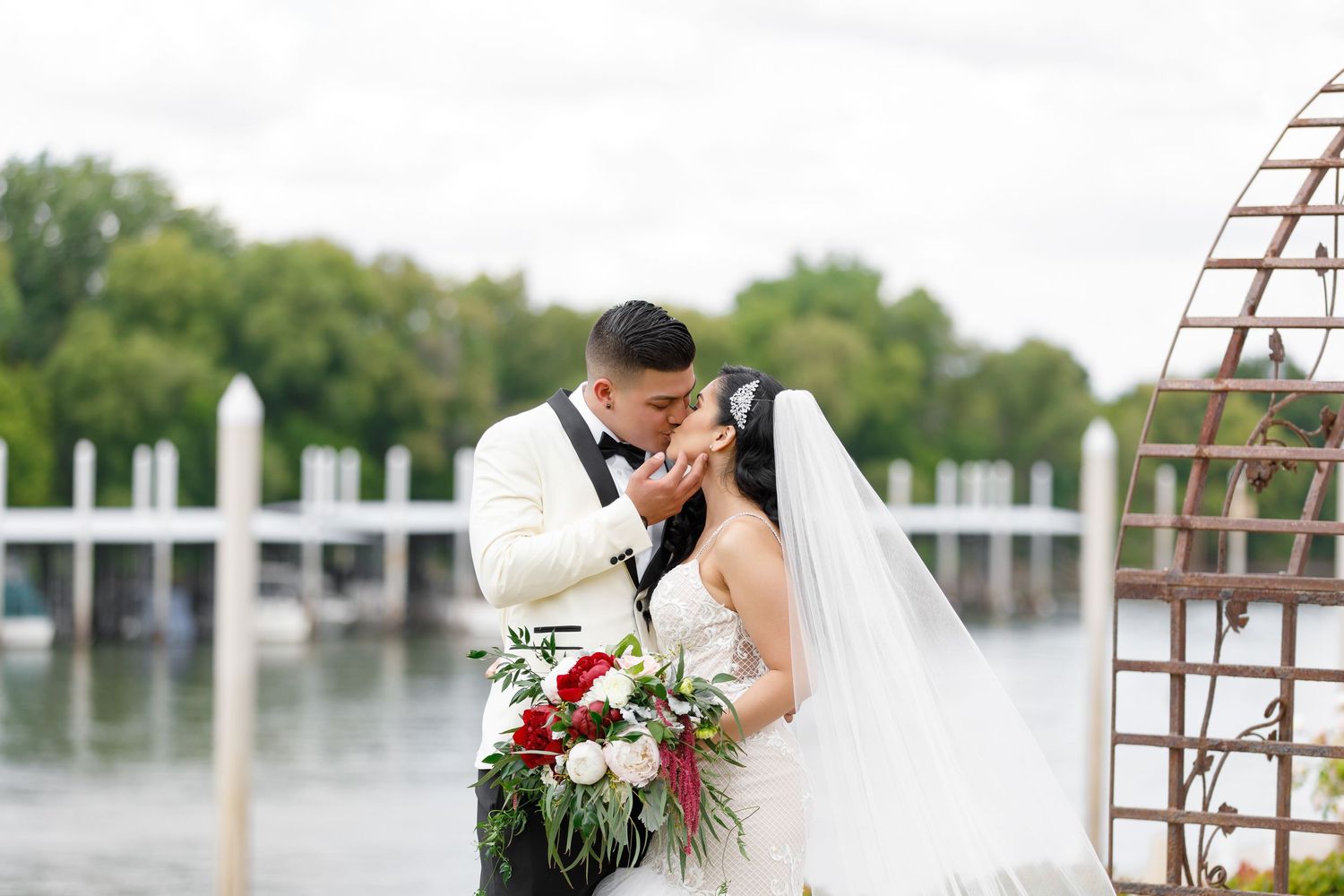 A couple shares a romantic moment in front of a waterfront setting with boats and docks visible in the background.