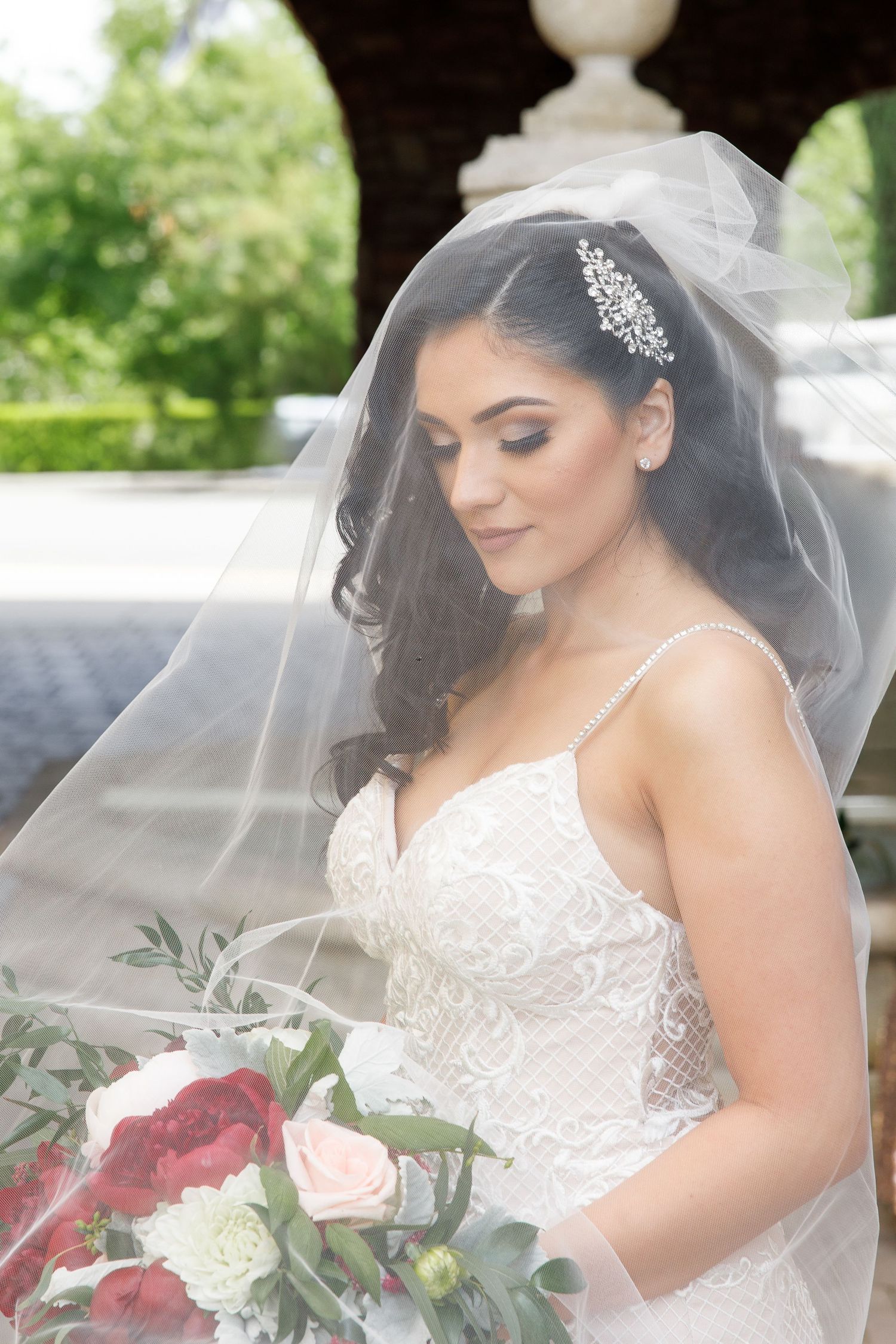 Bride in white lace wedding dress and veil holds a red and white bouquet in an outdoor garden setting.