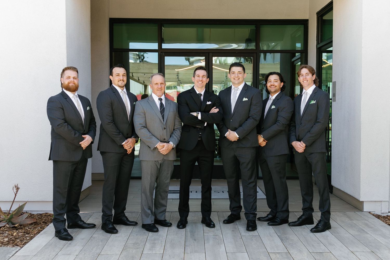 A group of groomsmen in black suits and ties stand together on a porch for a formal wedding photo.