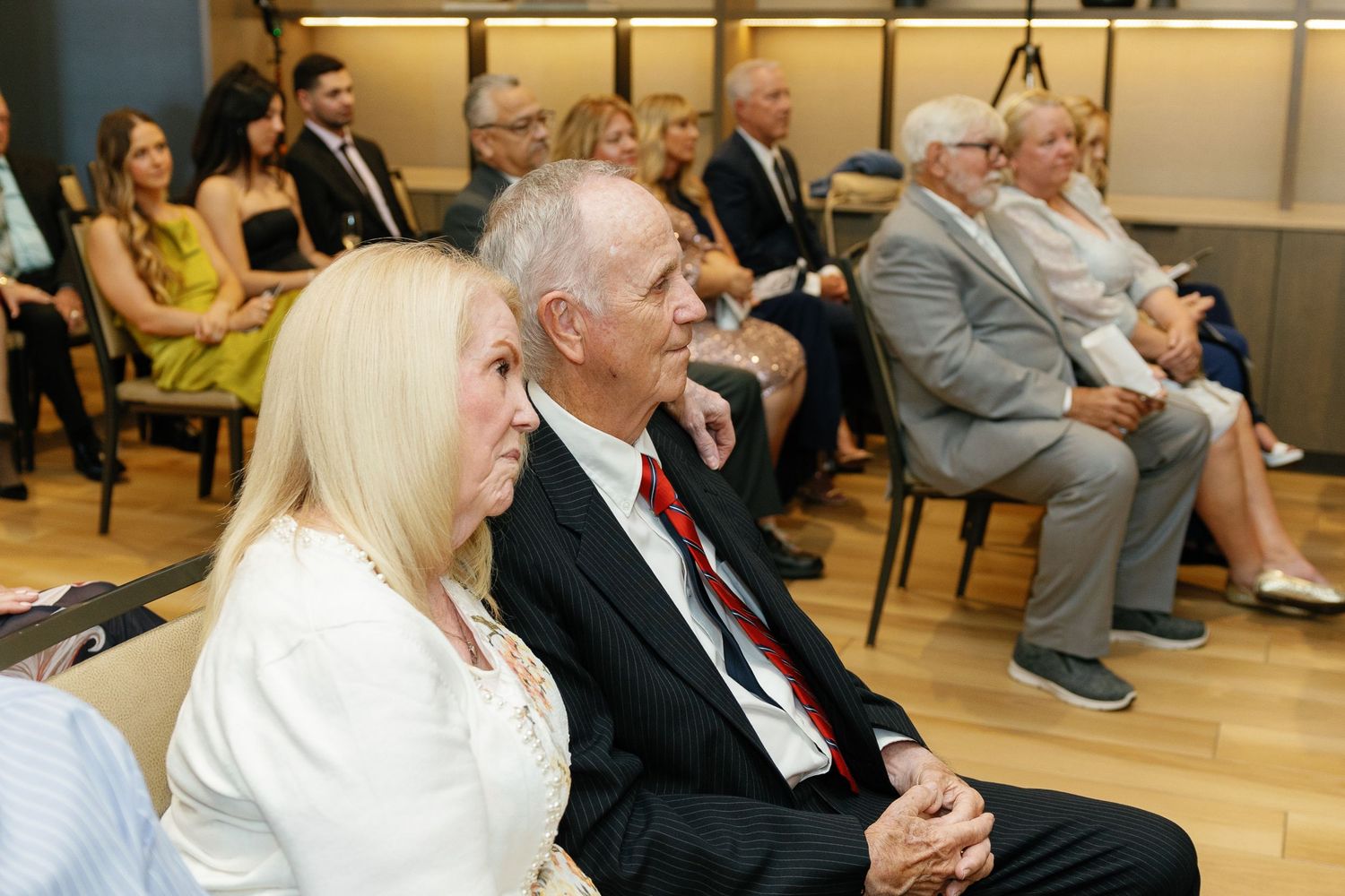 People seated in rows at an indoor event or meeting with warm lighting and a modern interior setting.