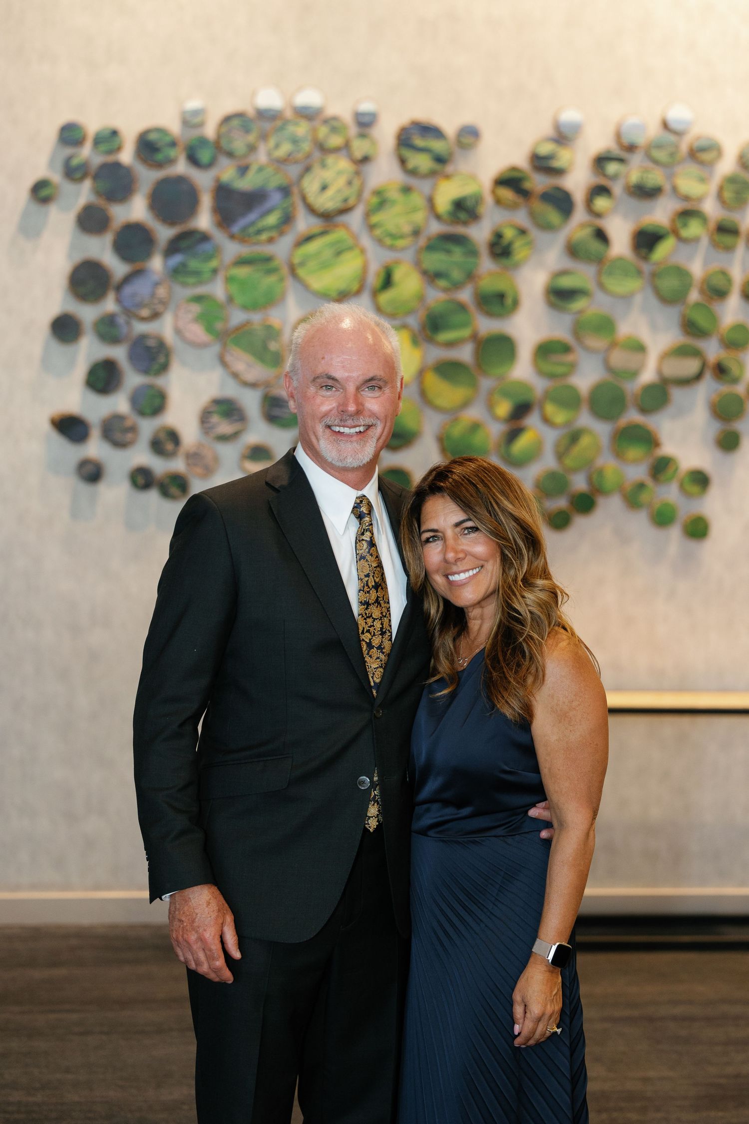 A couple in formal attire smiles together against a decorative wall with circular green art pieces.