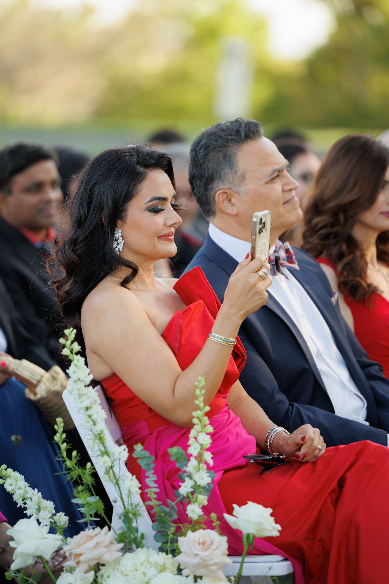 Wedding guests in formal attire sit in rows during an outdoor ceremony with white floral decorations.