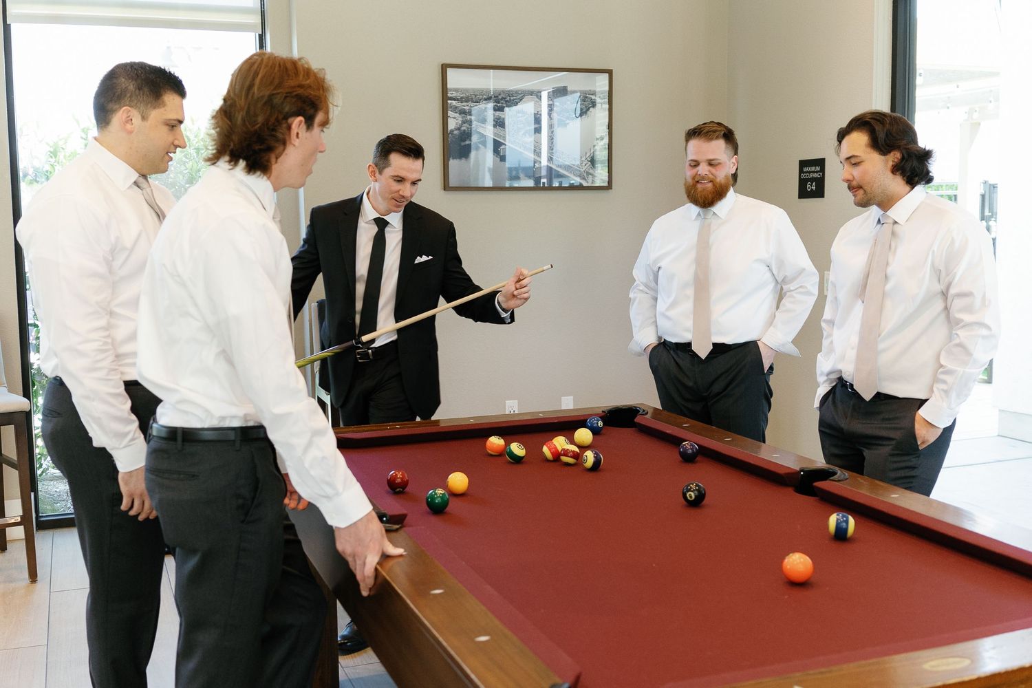 Well-dressed groomsmen in white shirts gather around a red pool table during a wedding day celebration.
