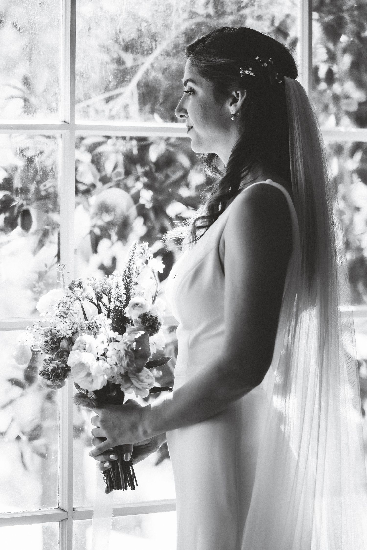 Black and white photo of a bride in an elegant sleeveless gown holding a white floral bouquet by a window.