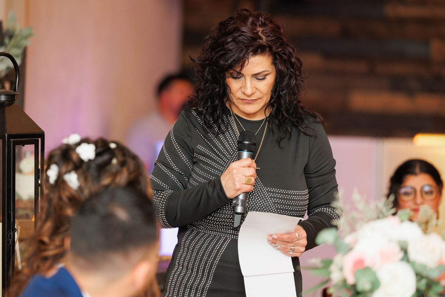 A speaker in a black striped sweater reads from a paper during an indoor event with soft pink lighting and floral decor.