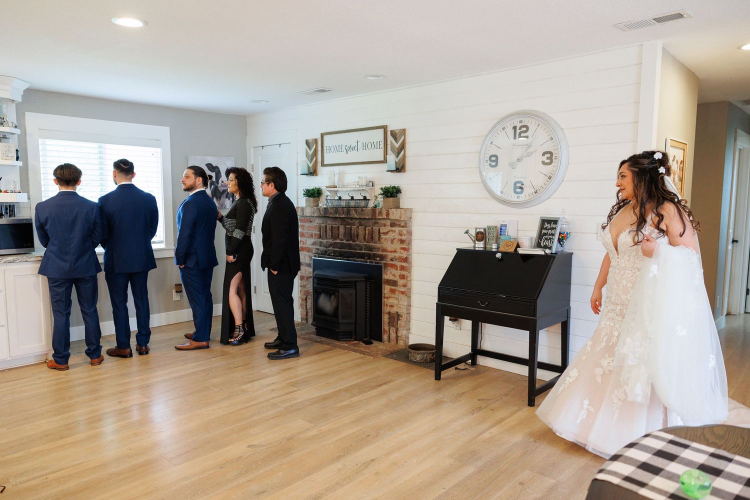 Wedding party gathers in a bright living room with hardwood floors, white walls, and a decorative piano and clock.