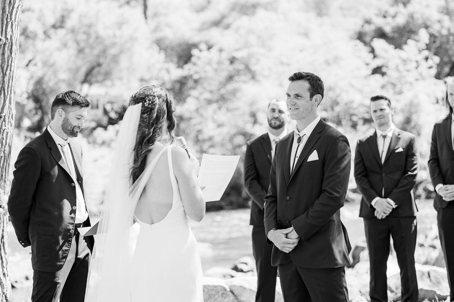 Black and white photo of an outdoor wedding ceremony with the couple and wedding party gathered in a natural setting.