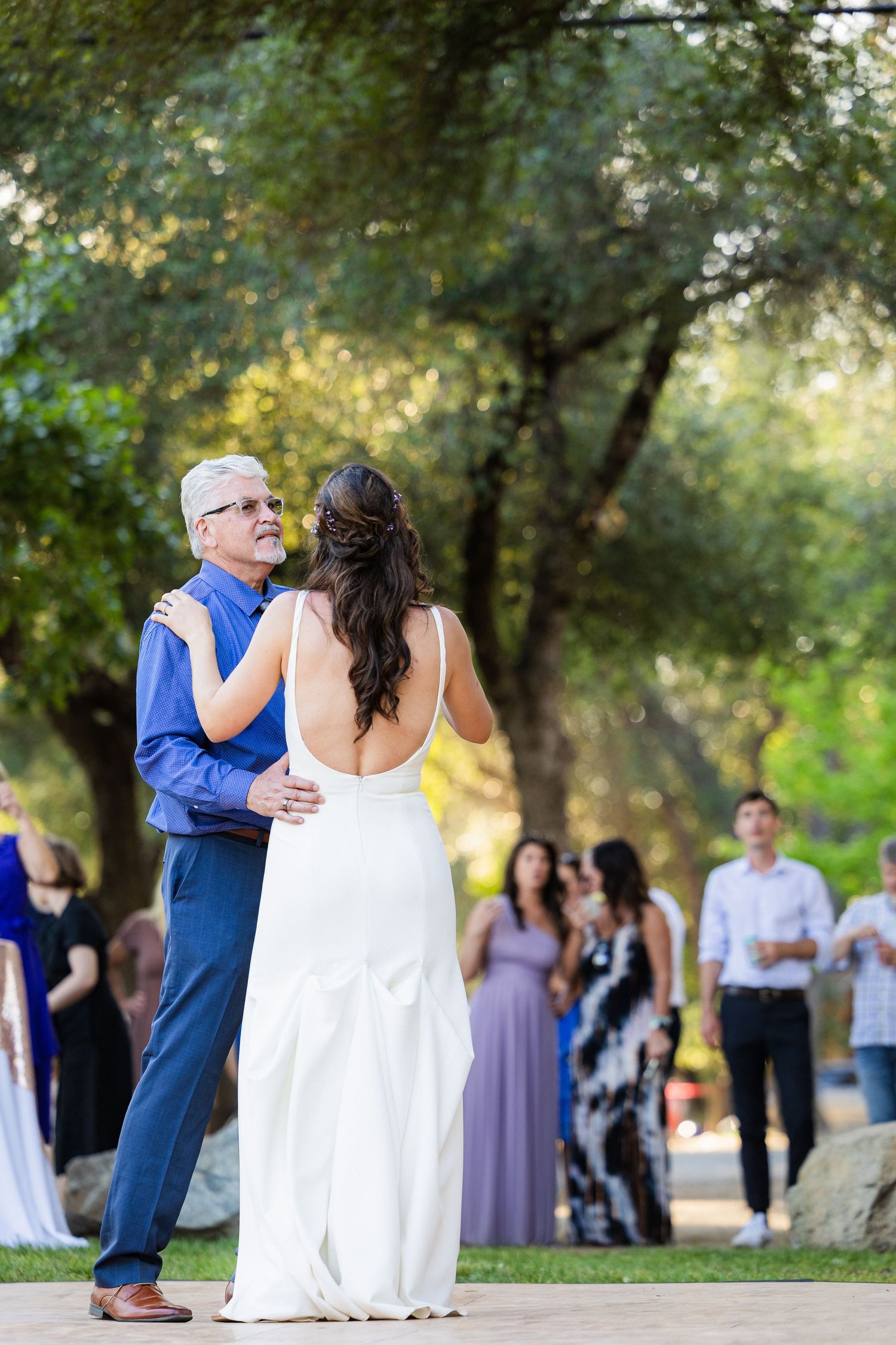 A couple shares their first dance together at an outdoor wedding reception surrounded by trees and guests.