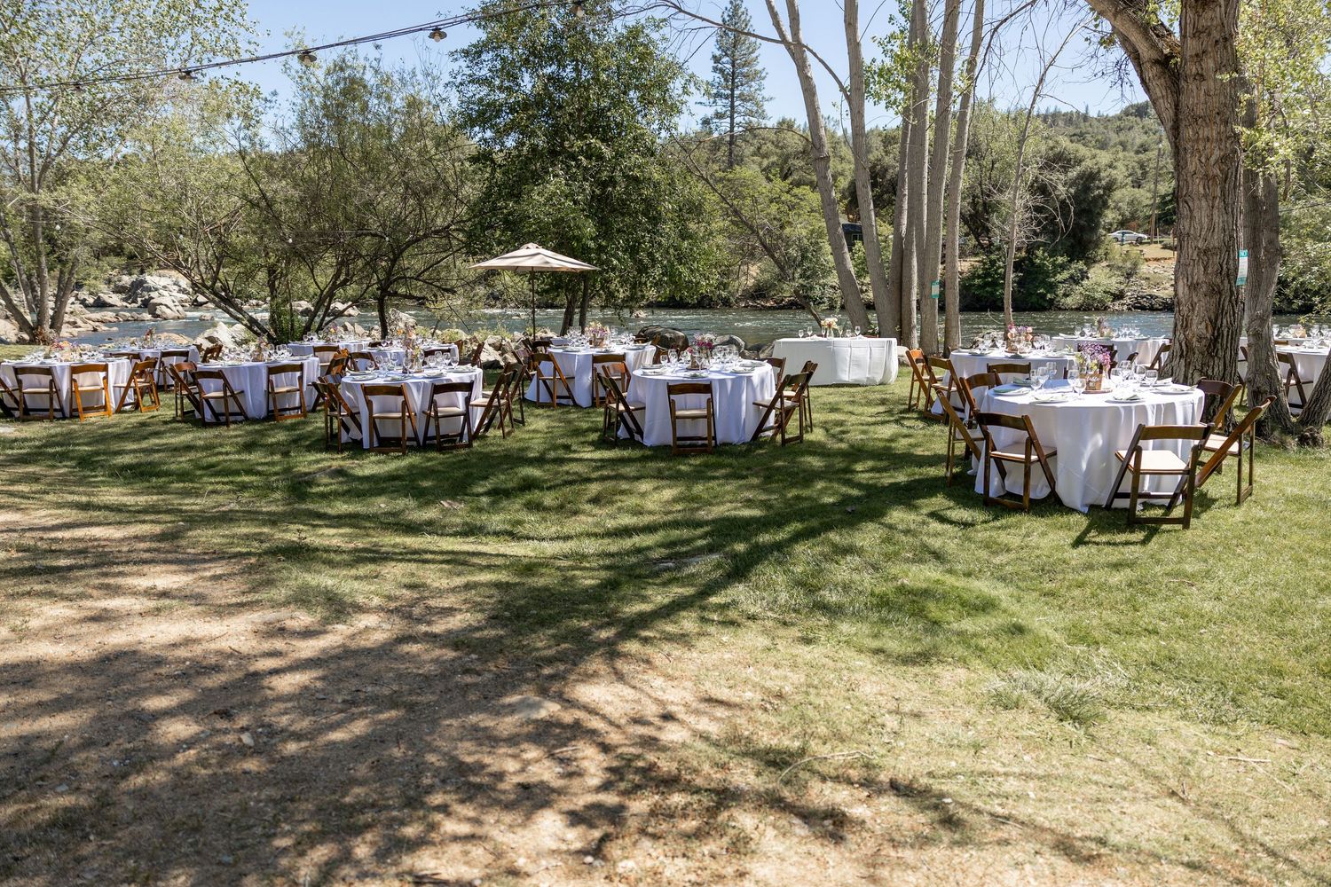 Tables and chairs arranged under trees at an outdoor garden venue with string lights and umbrellas for dining.