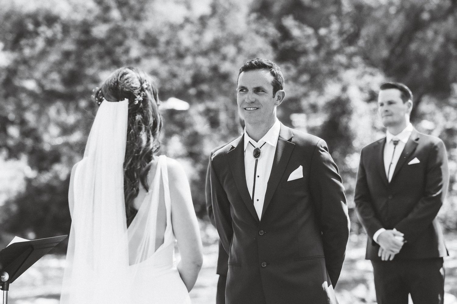 A black and white outdoor wedding ceremony photo showing a bride and groom exchanging vows with an officiant in the background.