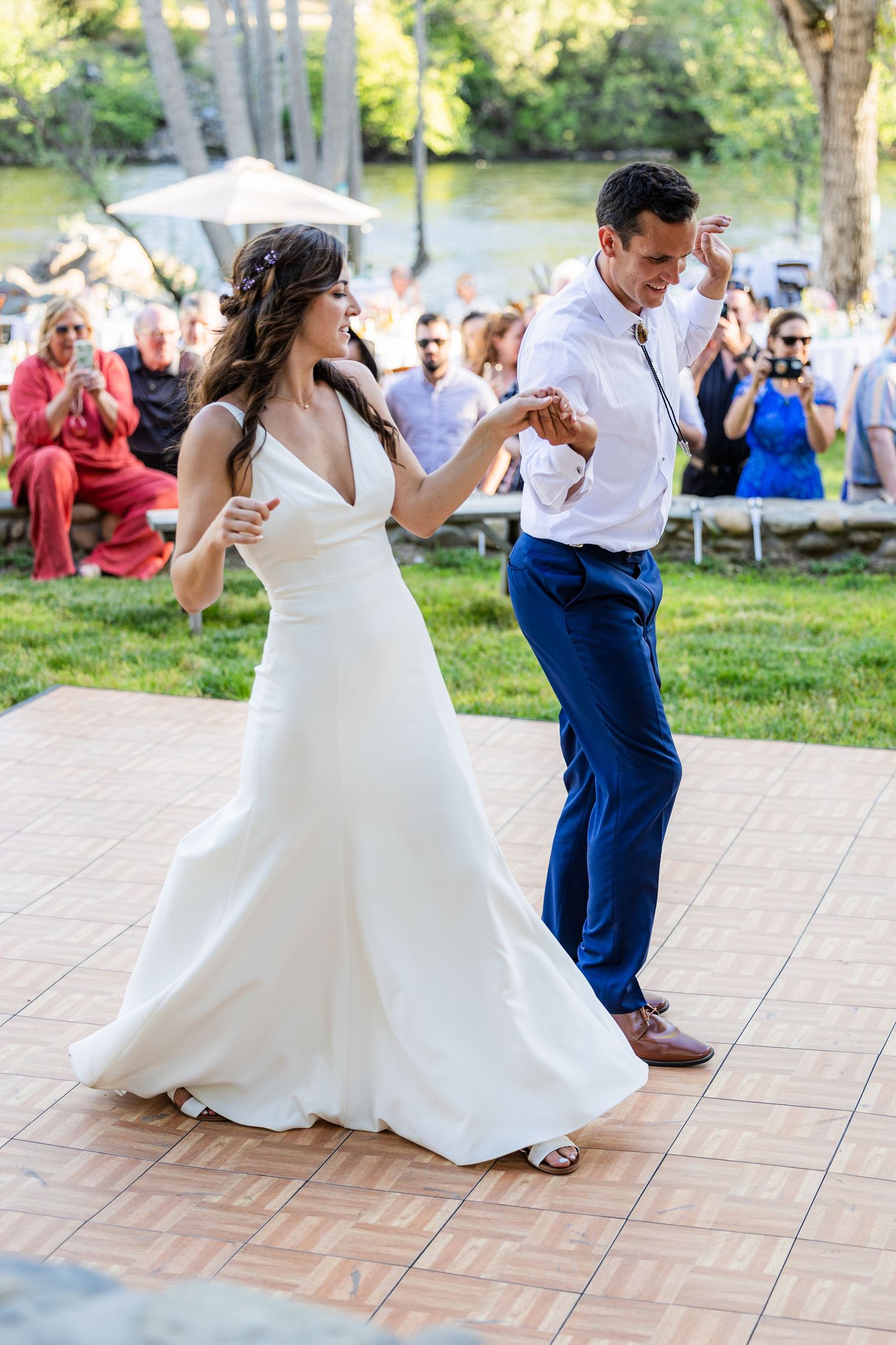 A couple dances together at an outdoor wedding reception on a tiled dance floor surrounded by trees and guests.