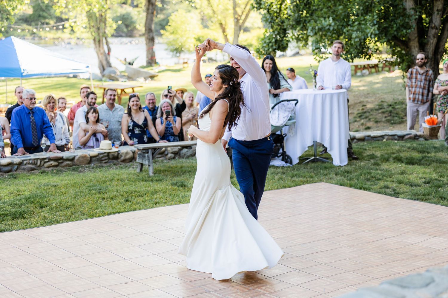 A couple shares their first dance on an outdoor wooden dance floor surrounded by wedding guests in a garden setting.