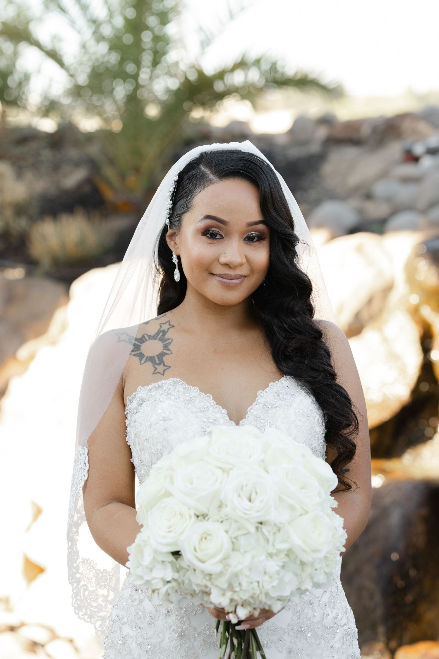 Beautiful bride in lace wedding dress holds elegant white rose bouquet against natural outdoor backdrop.