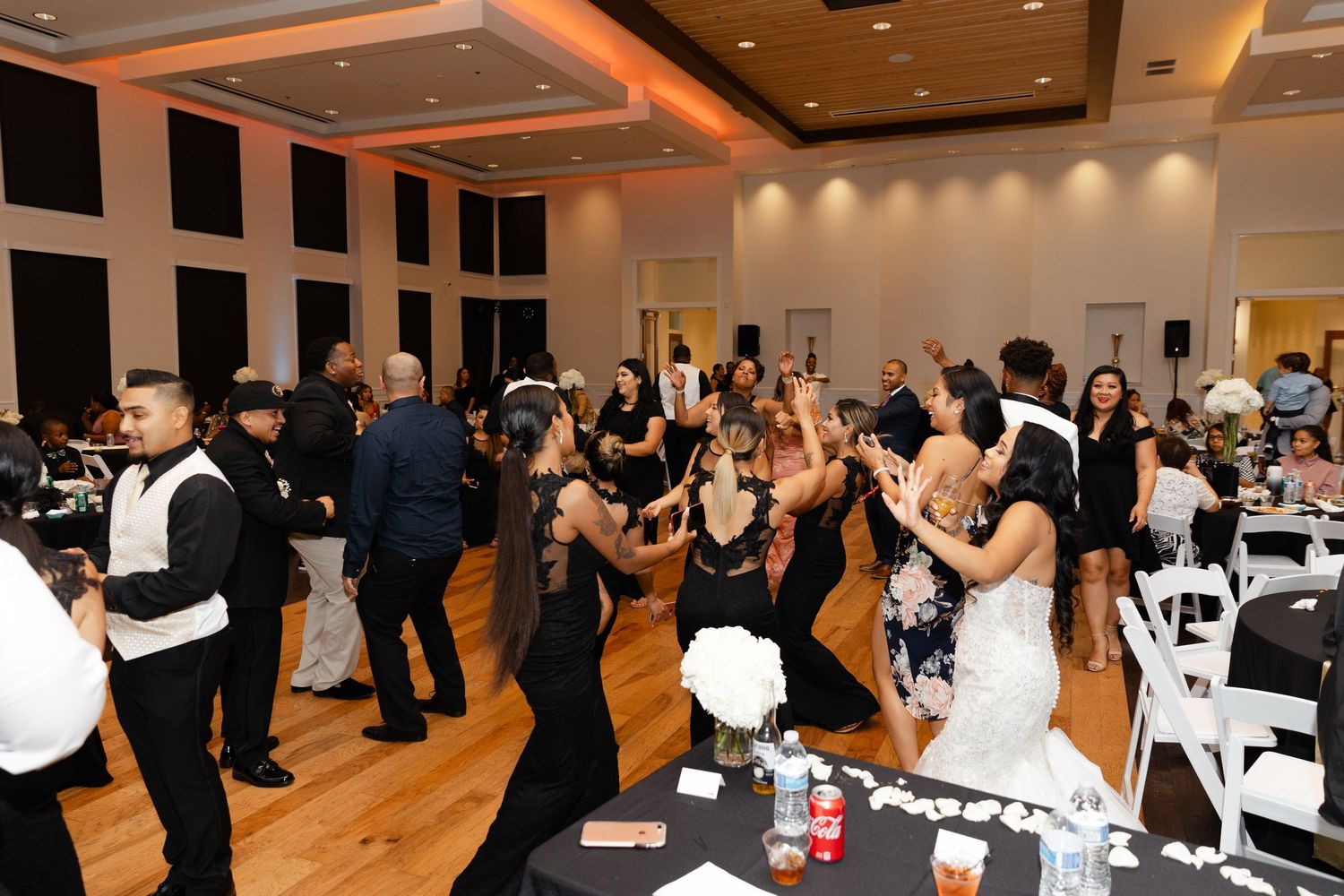 Wedding guests dancing and mingling at an elegant reception venue with warm lighting and white chair decor.