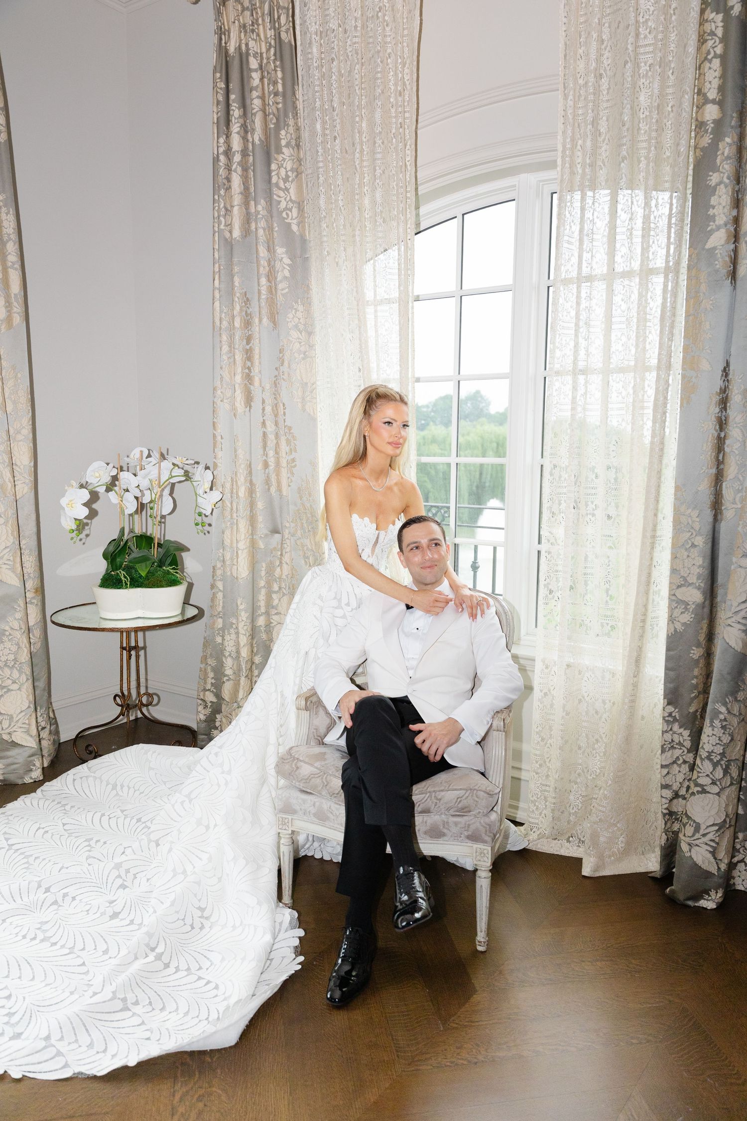 A couple in formal wedding attire poses playfully in a bright room with sheer curtains and elegant decor.