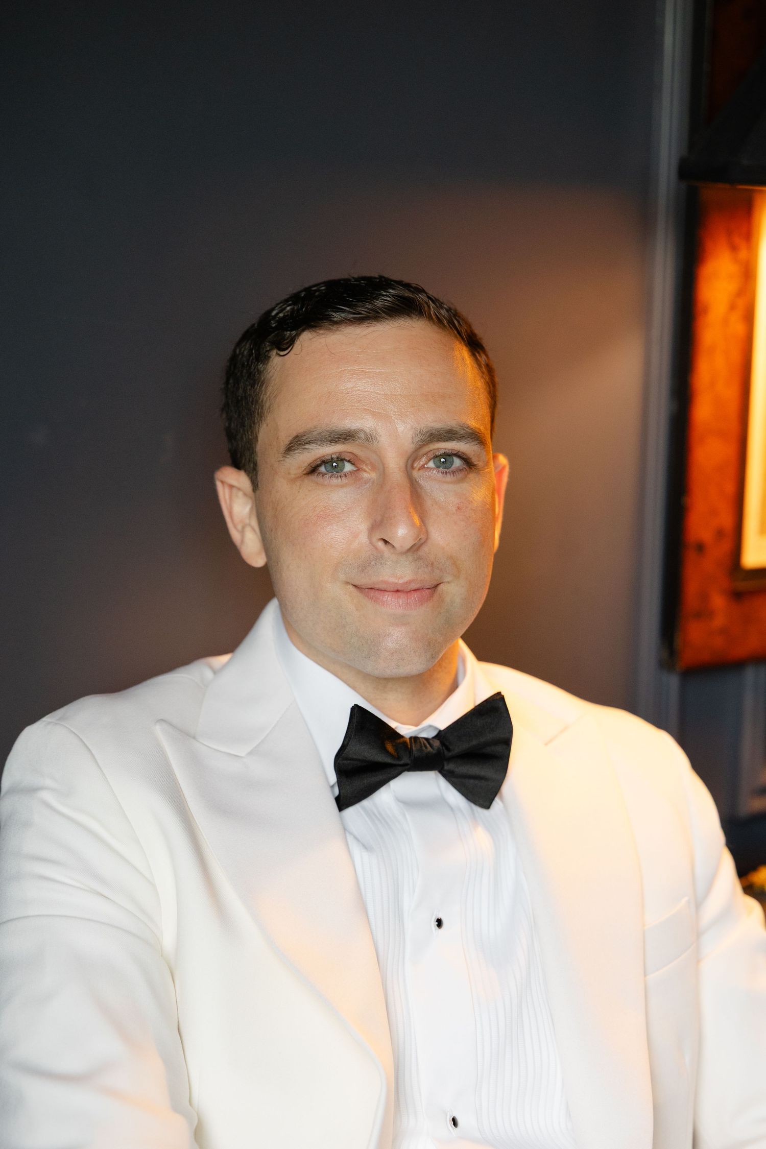 A person wearing a white tuxedo jacket and black bow tie smiles at a formal event.