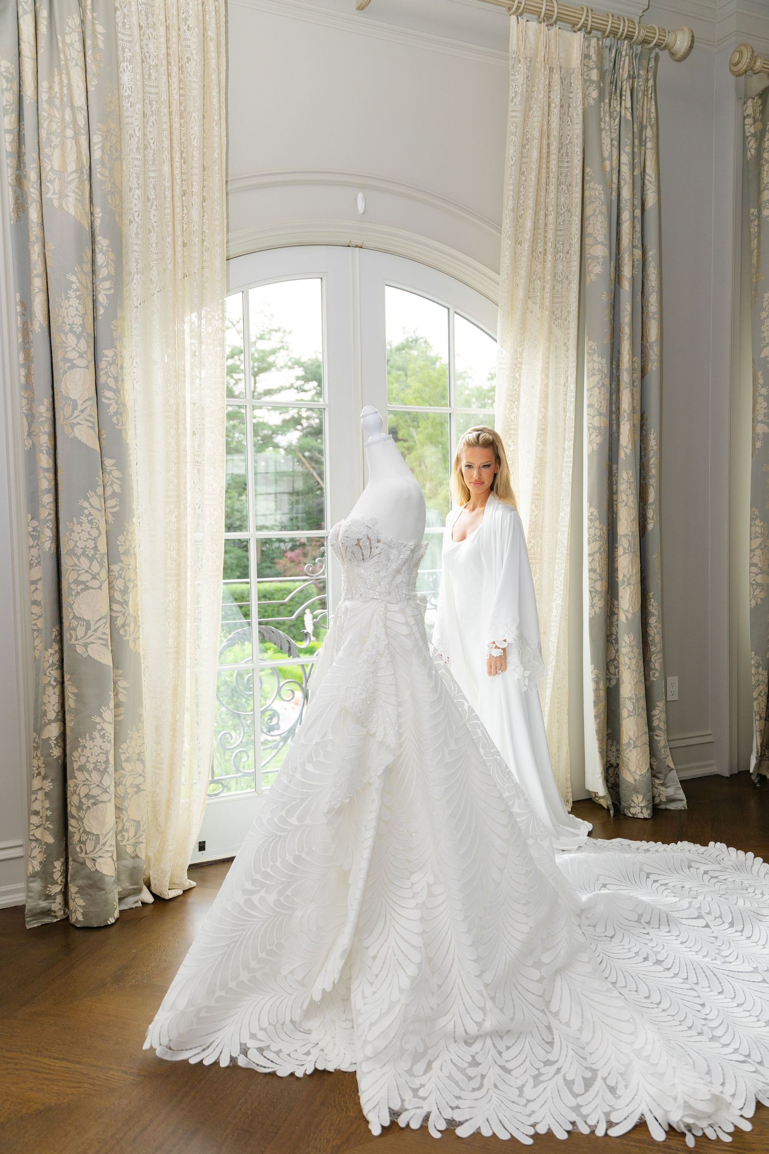 An elegant white lace wedding gown with long train displayed by arched window with flowing cream curtains.