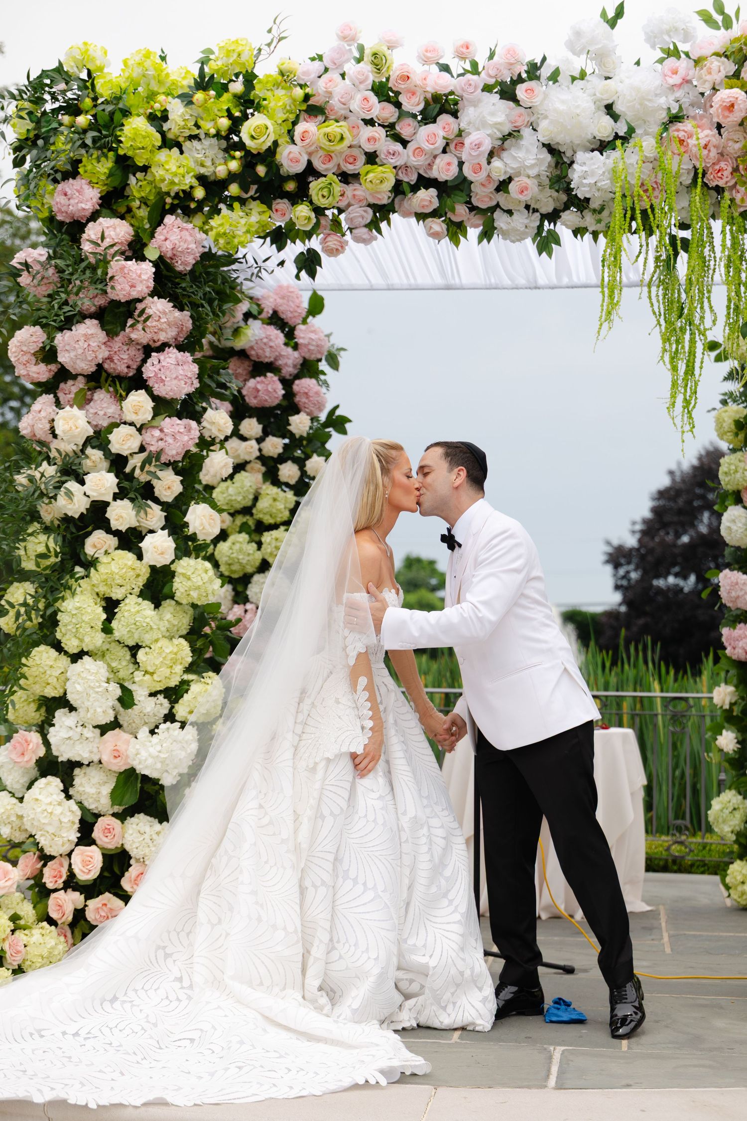 Romantic wedding couple embraces under an elegant floral arch with pink and white roses and cascading greenery.