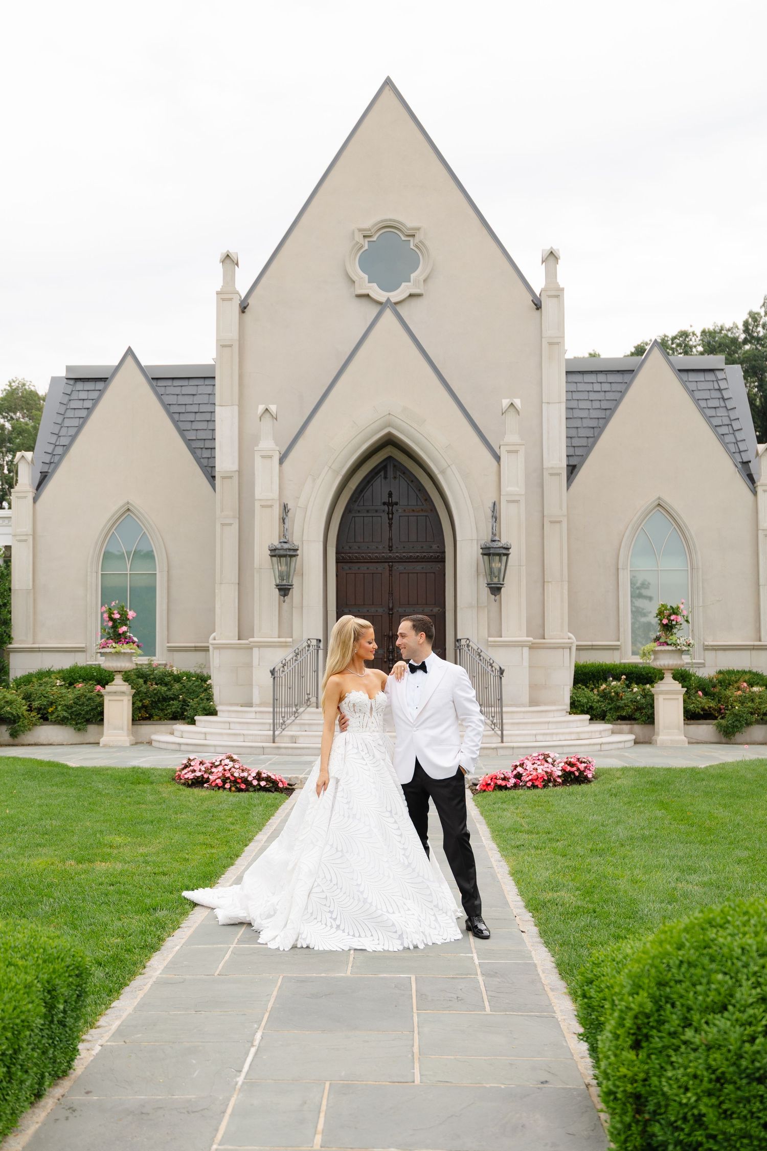 A wedding couple stands outside a beautiful gothic-style church with cream colored walls and arched windows.