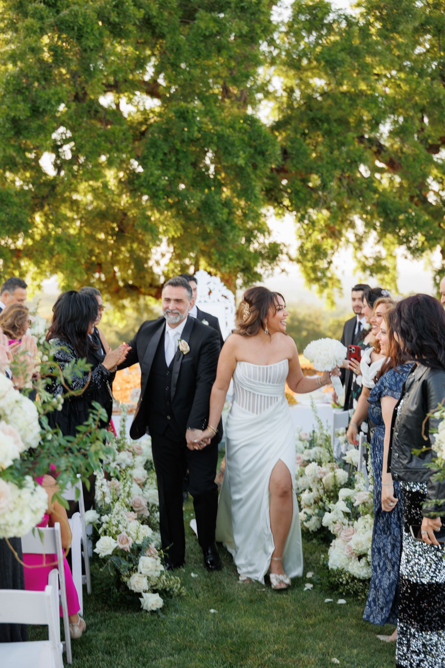 Wedding guests mingle and socialize during an outdoor garden reception with white floral arrangements at sunset.