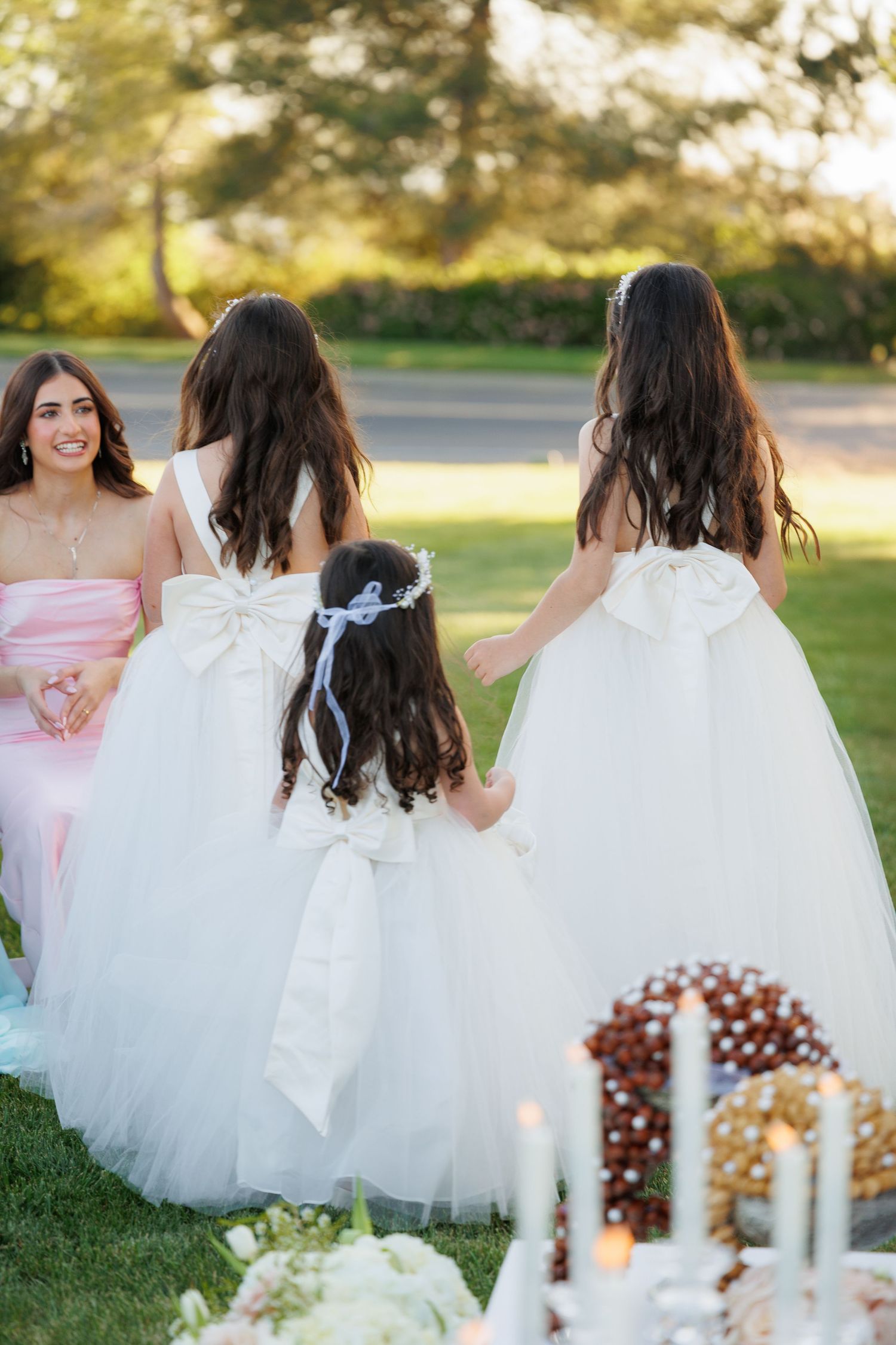 Little girls in white dresses sit together at an outdoor tea party with candles and decorations at sunset.
