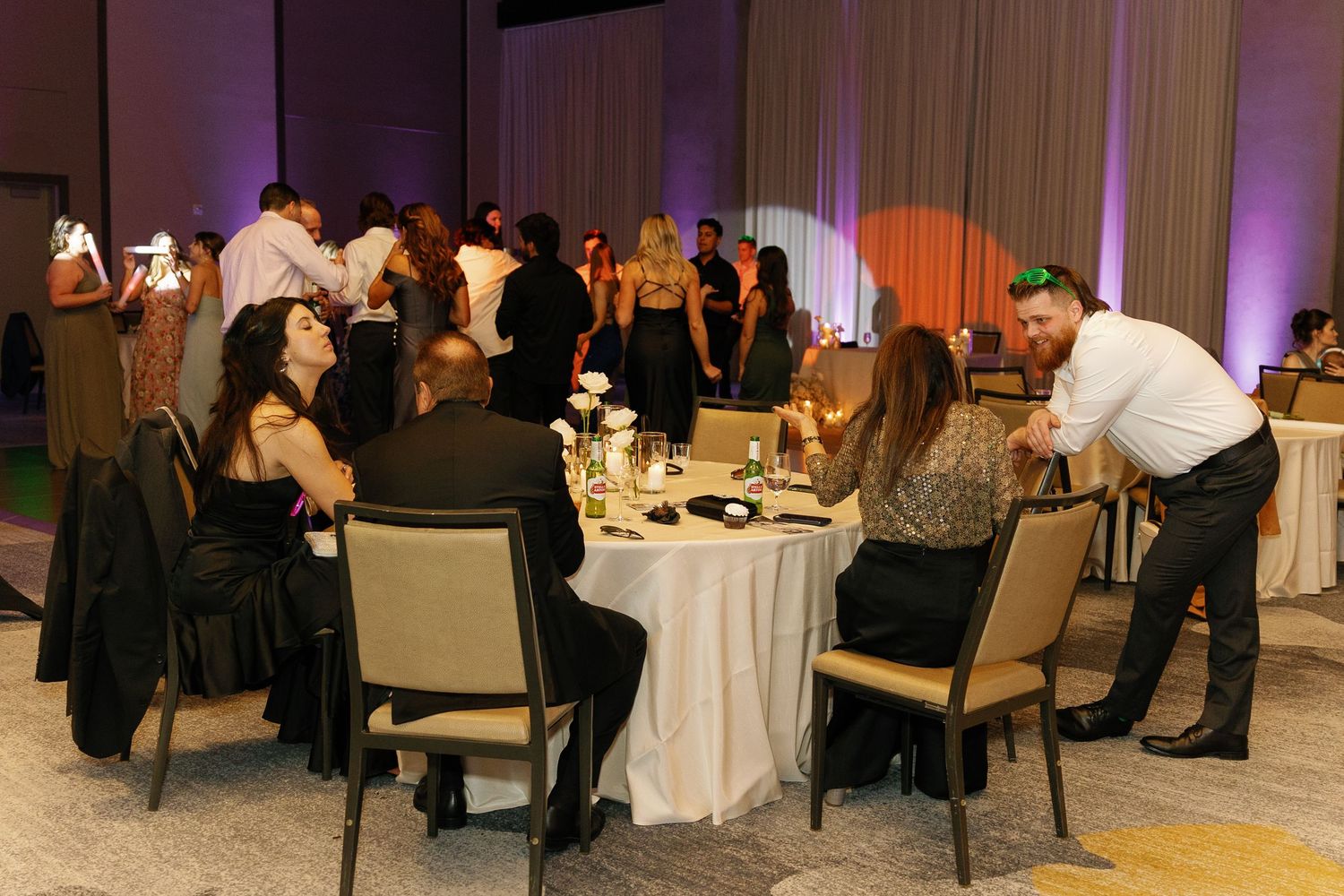Guests gather around elegant tables at an indoor wedding reception with purple uplighting and white linens.