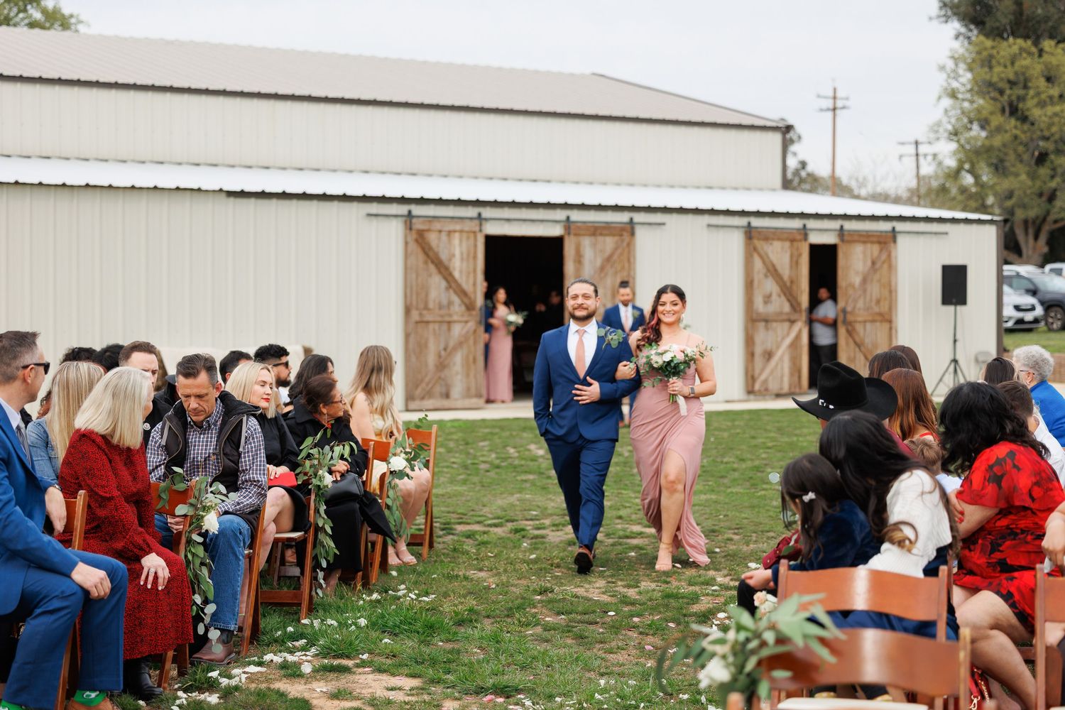 Wedding party members walk down the aisle between seated guests at an outdoor barn ceremony.