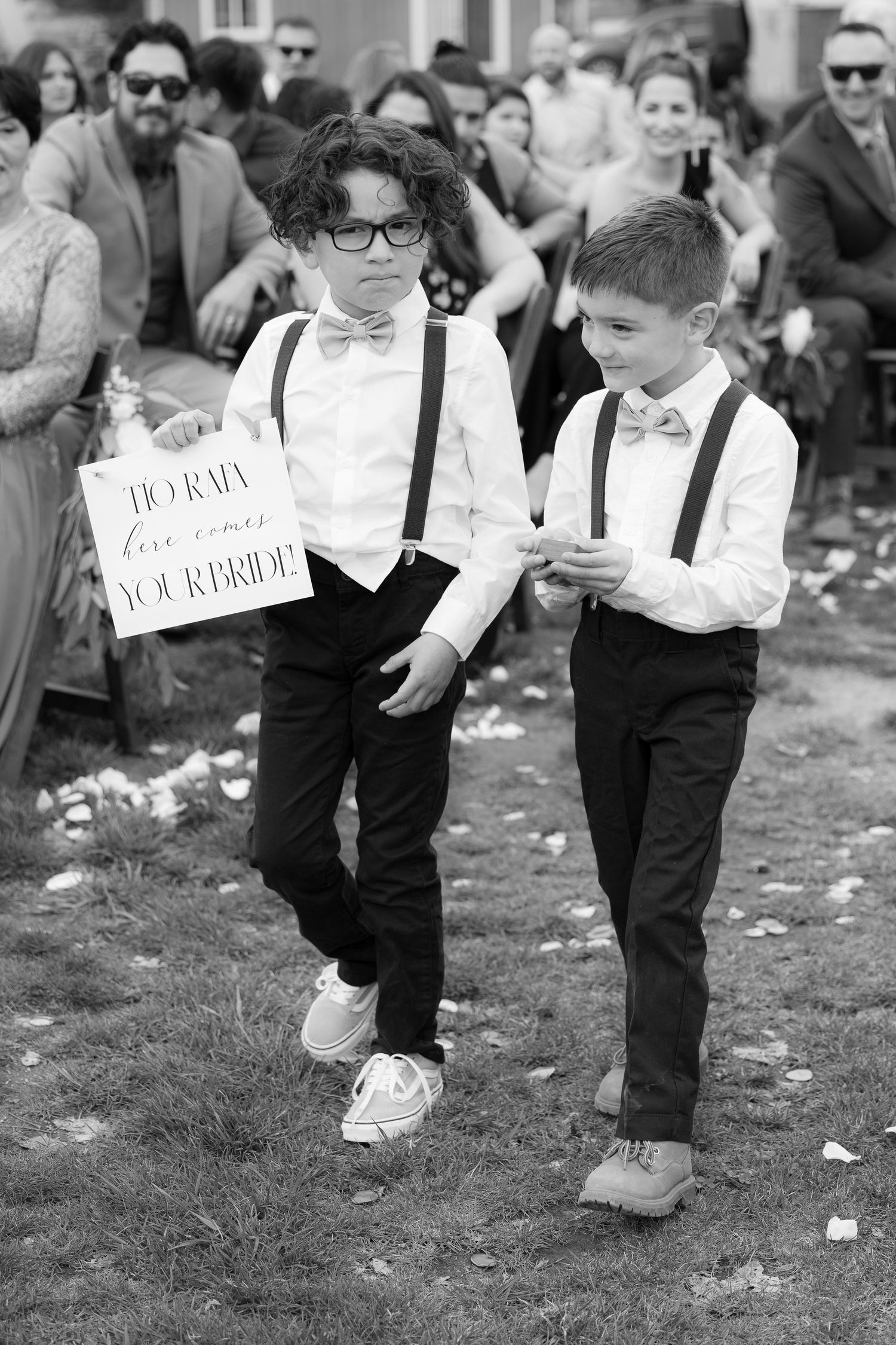 Two young ring bearers in bow ties and suspenders walk together at a wedding ceremony, one holding a sign.