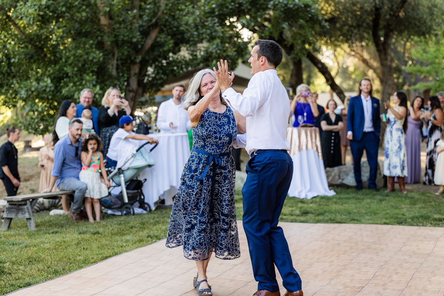 Guests enjoying an outdoor wedding celebration with dancing on a lawn surrounded by trees.