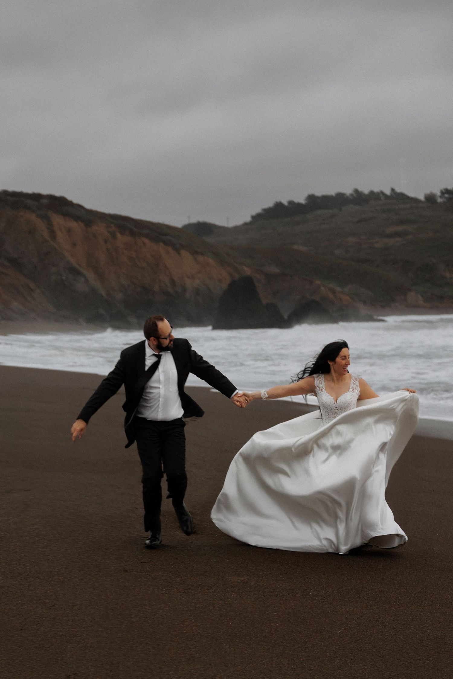 A couple in wedding attire plays dramatically on a moody beach with dark sand and cliffs in the background.