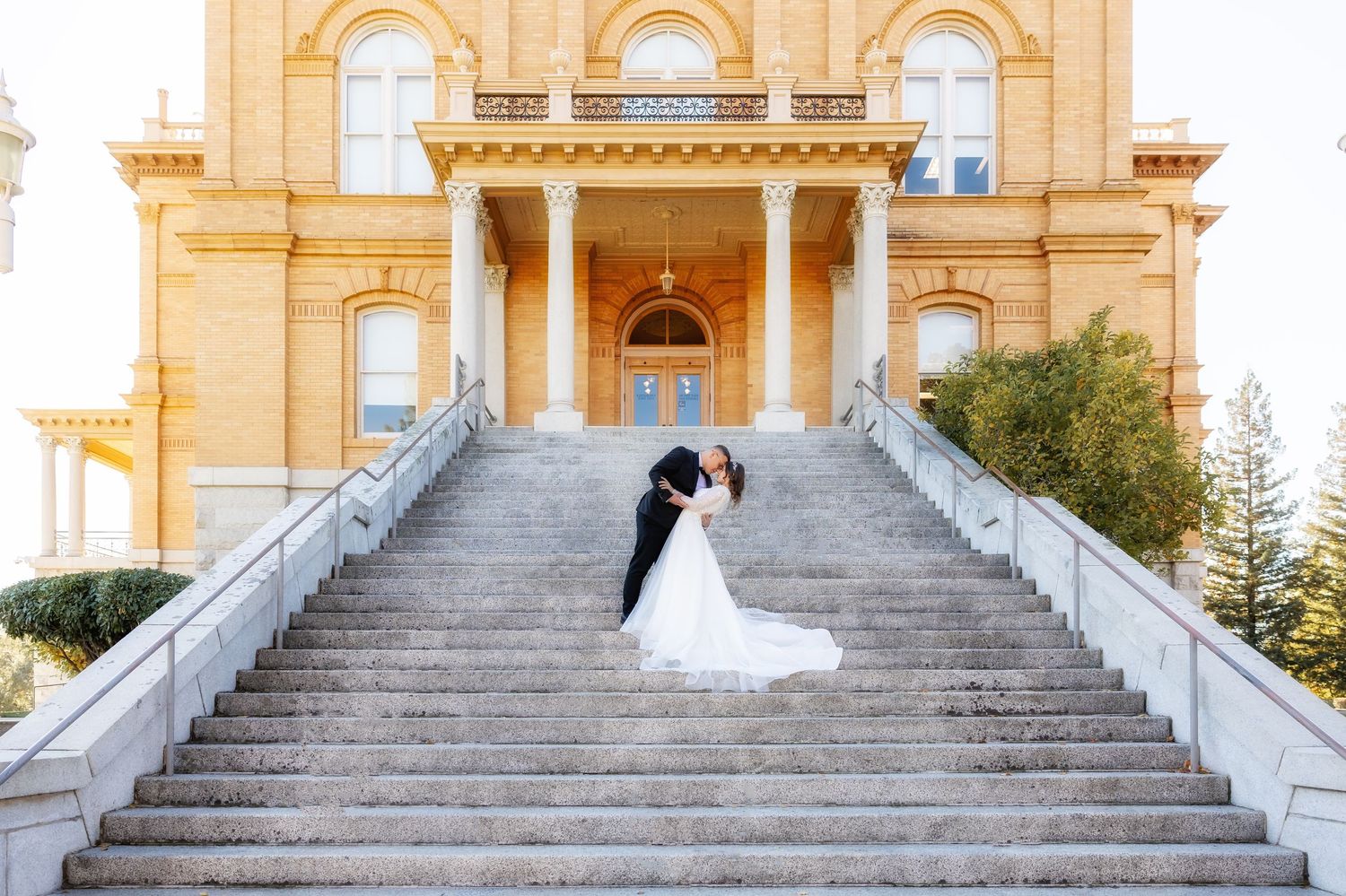 Wedding couple poses on grand stone staircase of historic yellow building with arched windows.