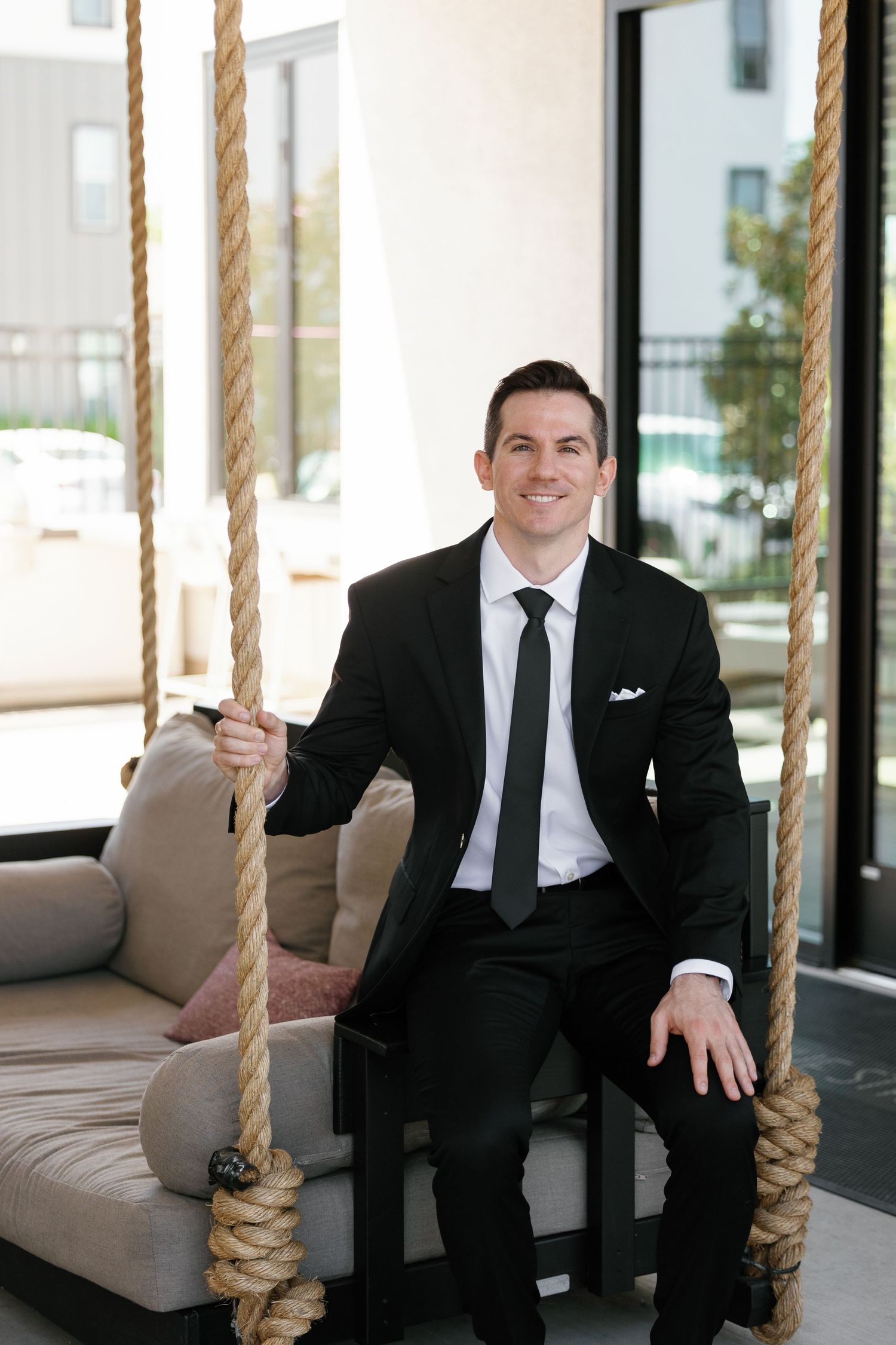 Professional in black suit and tie sits on an elegant hanging porch swing with rope supports.