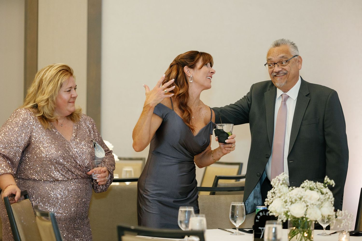 Three people share an animated moment together at a formal event with white floral centerpieces on the table.