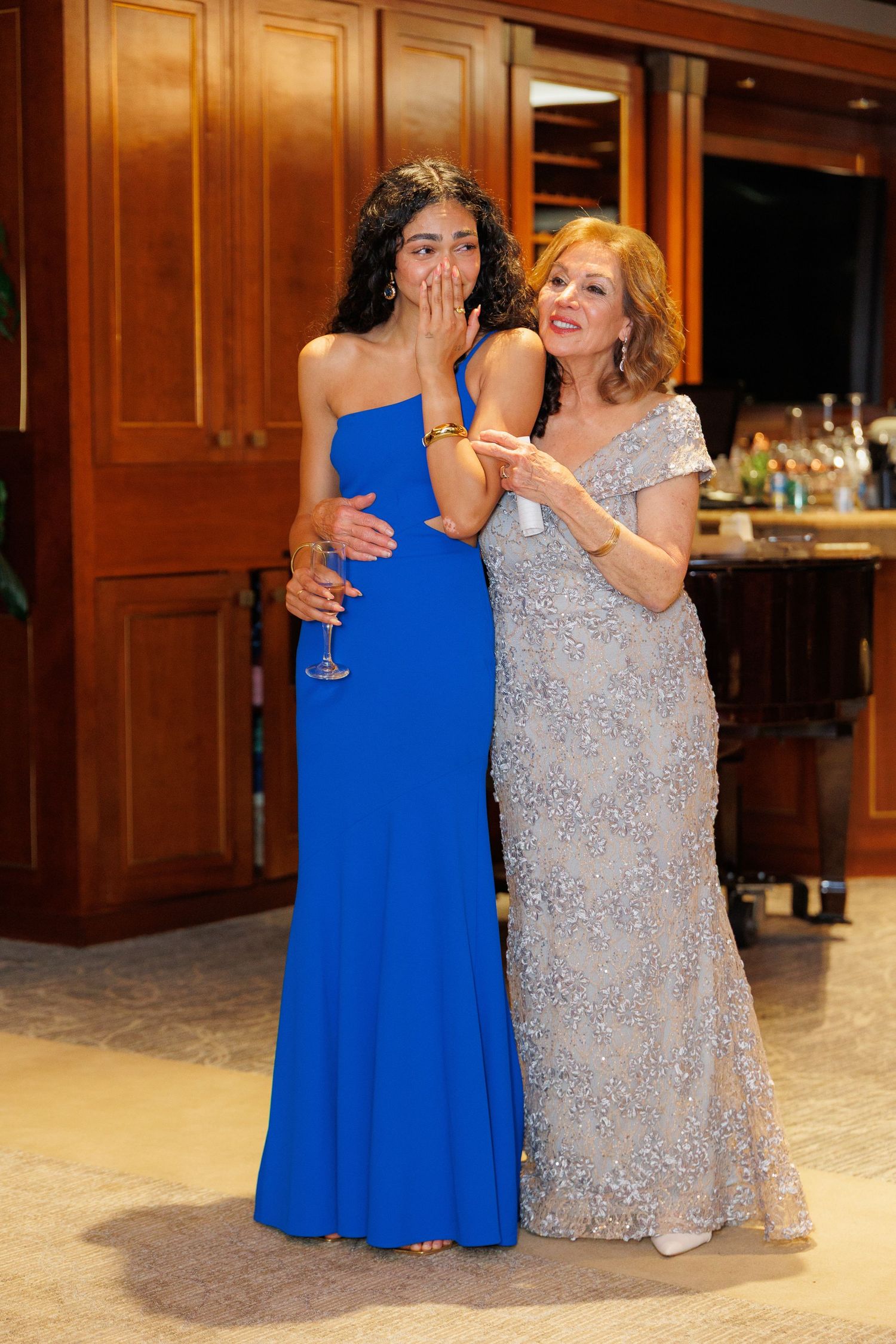 Two women in formal gowns, one in royal blue and one in silver sequins, share an emotional moment at an event.