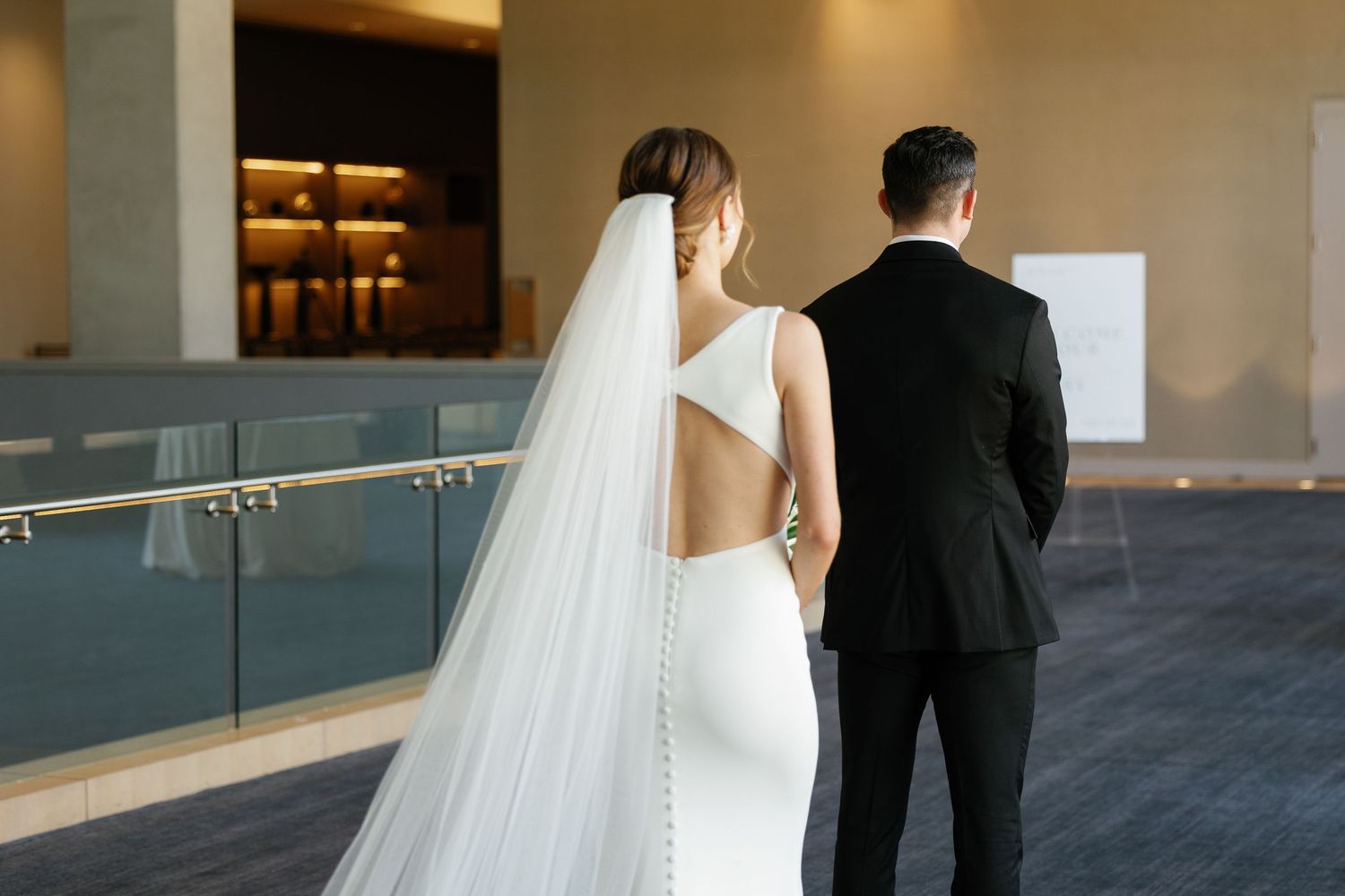 Couple in wedding attire stands together in an elegant indoor venue with glass walls.
