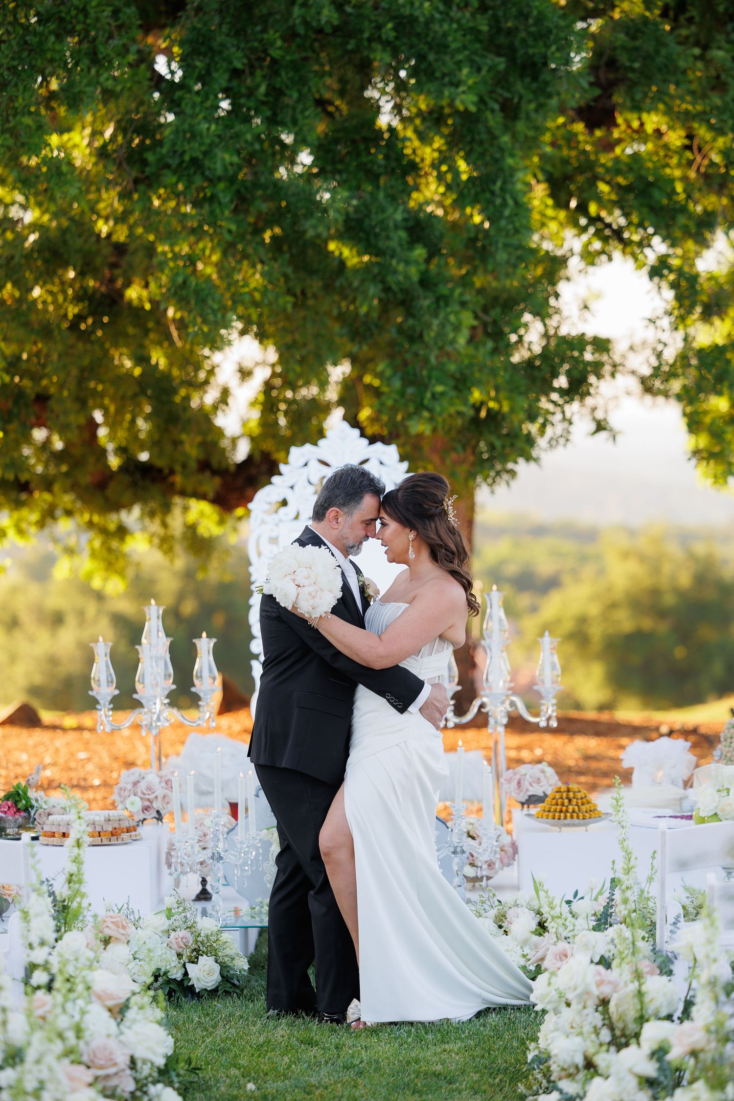 Romantic wedding scene with white floral arrangements and table settings under a tree at golden hour.